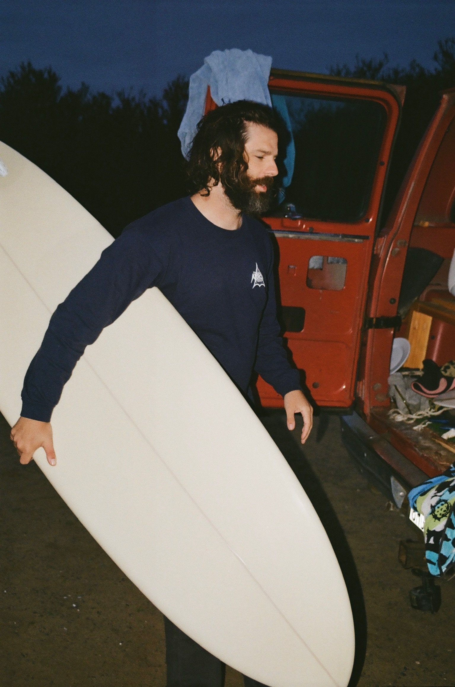 A man with dark, curly hair and a beard holding a white surfboard, standing next to an orange utility vehicle with open door, nighttime setting.