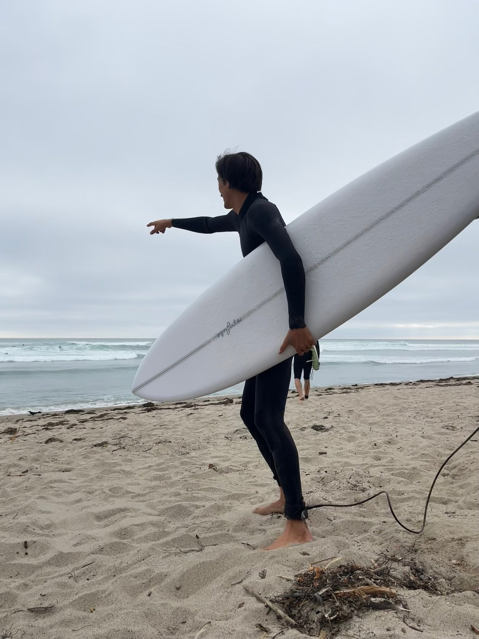 A person in a wetsuit holding a surfboard on a sandy beach in San O, pointing towards the ocean with overcast skies.
