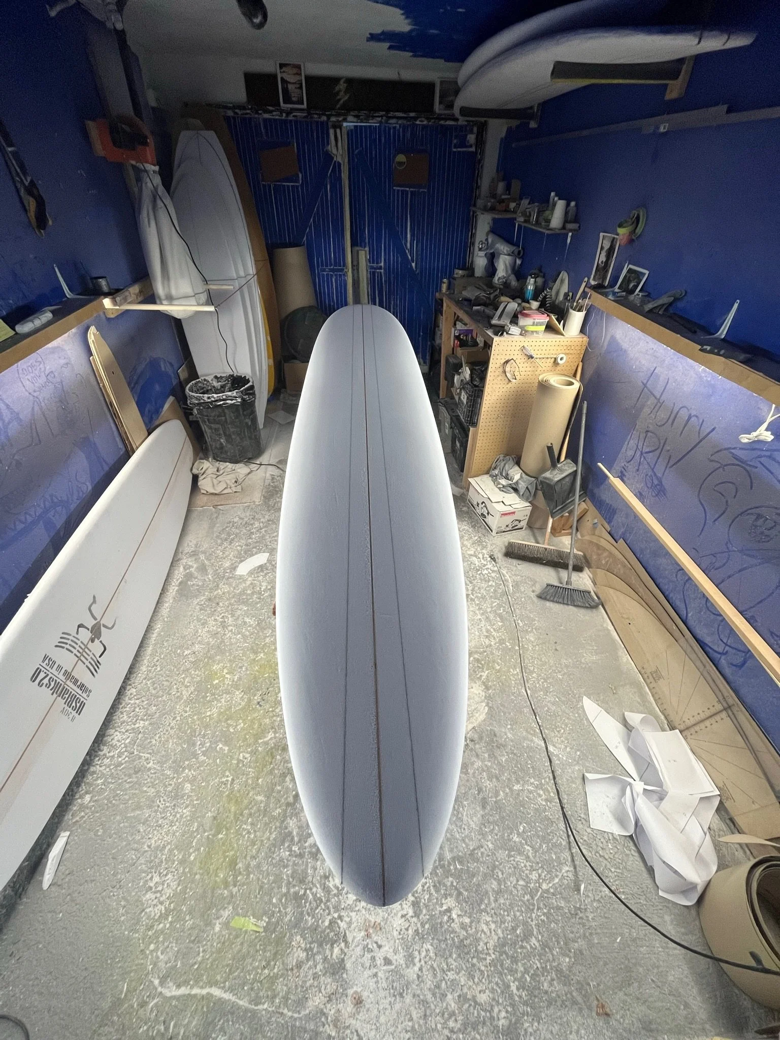 Angle of a surfboard in a workshop with tools, surfboards, and supplies on shelves and walls.