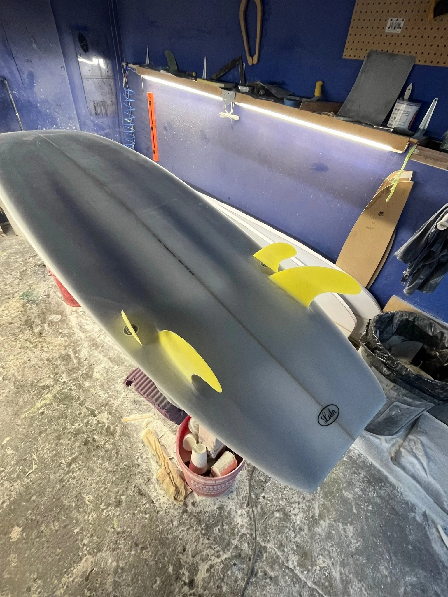 Indoor workshop with a square tail surfboard, tools, and supplies, including a workbench, pegboard, scissors, and storage containers.