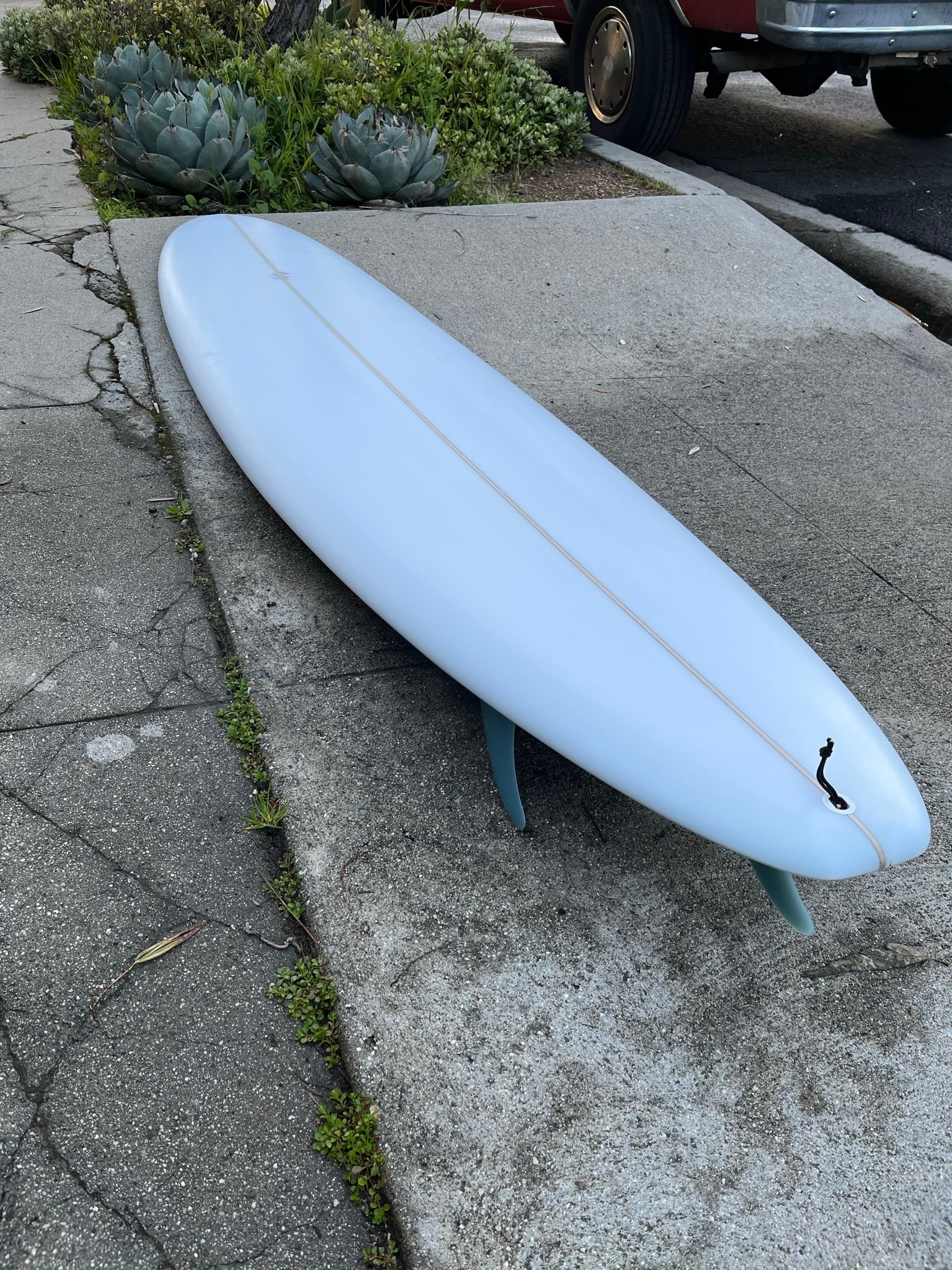A white surfboard lying on a cracked concrete sidewalk next to a small patch of greenery containing succulents and grass. A red vehicle is partially visible in the background.