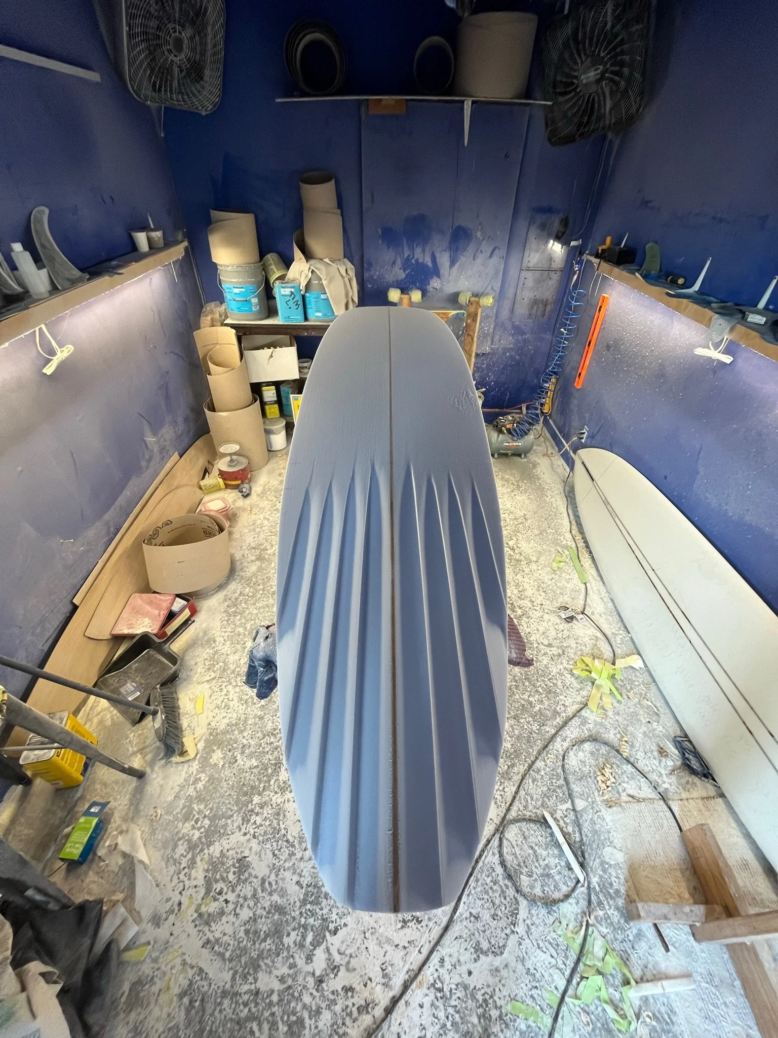Inside a workshop with blue walls, various tools, paint cans, and equipment scattered on shelves and the floor. A surfboard  with Channel Bottom is positioned vertically in the center, with work tools and supplies around.
