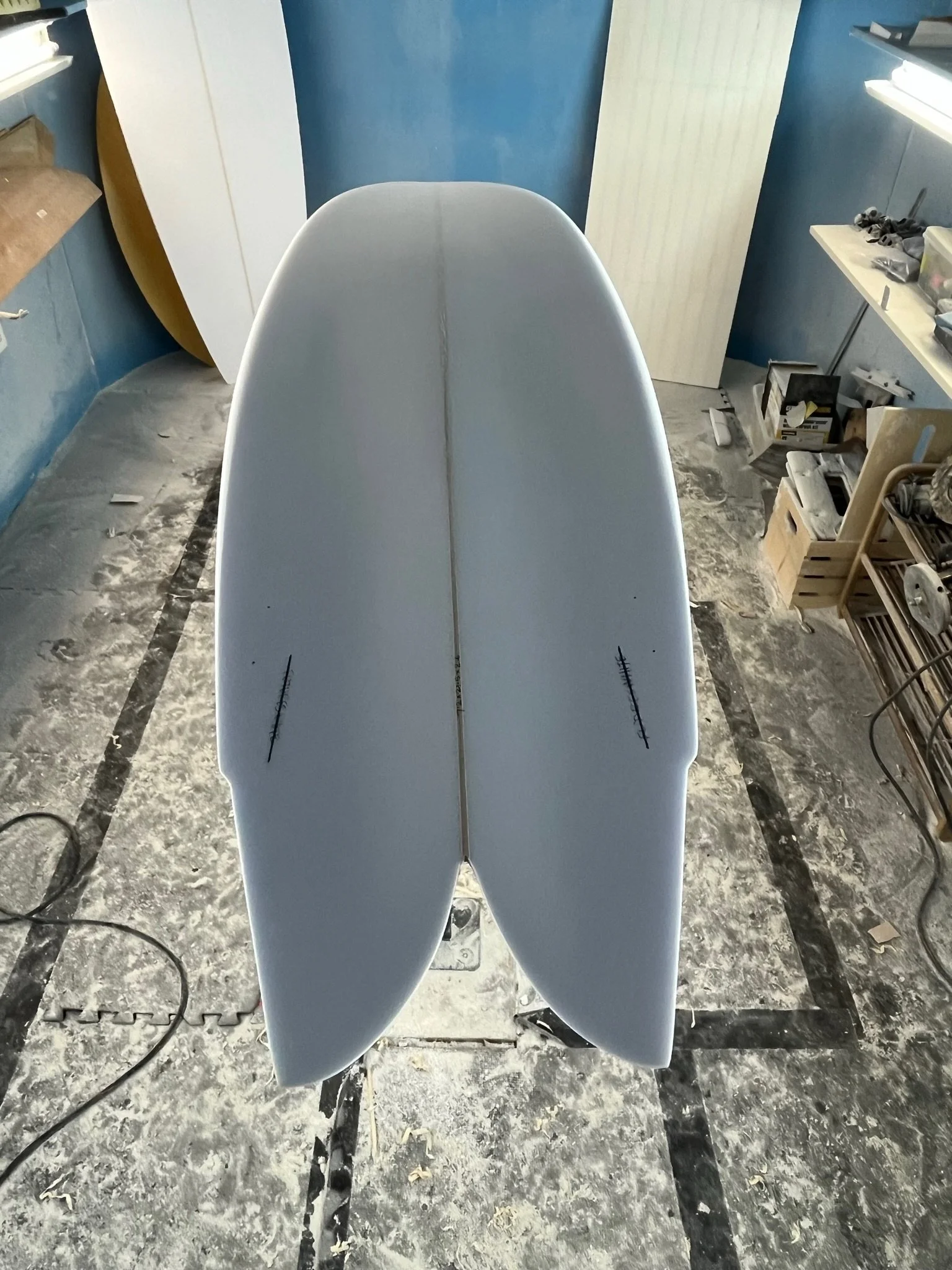 A surfboard is standing upright in a workshop or garage space, with various tools and materials around. The floor appears to have dust or powder on it, and there are shelves with items to the right.