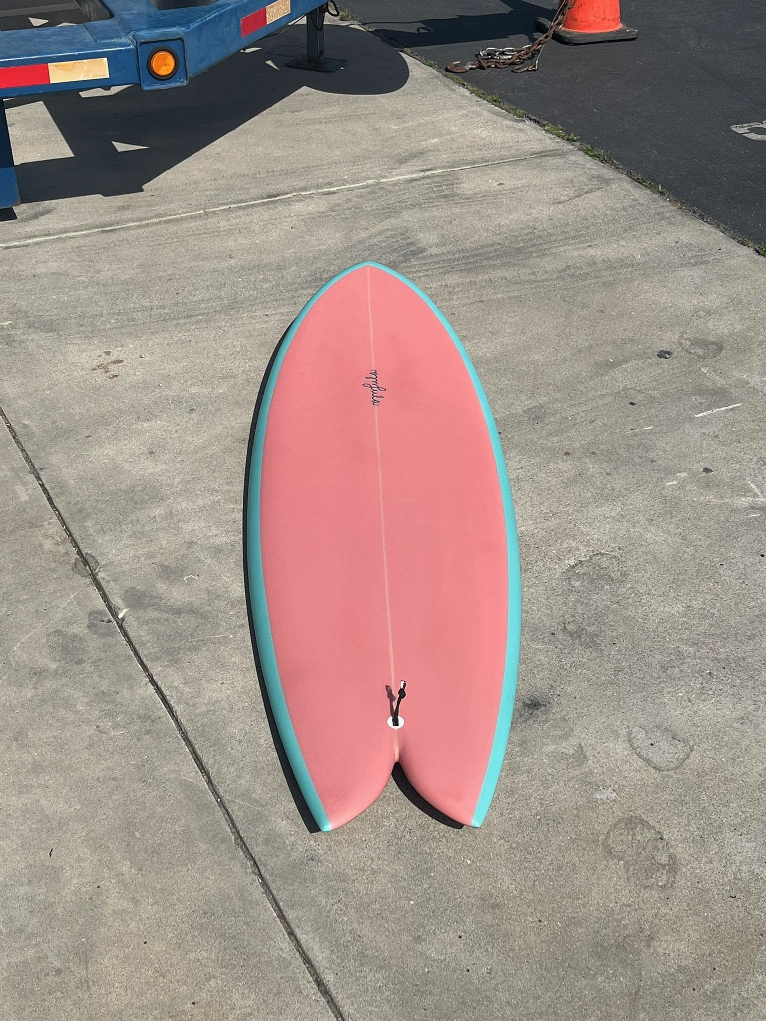 A pink stand-up paddleboard with turquoise edges, resting on a concrete sidewalk near a construction cone, chain, and vehicle trailer.