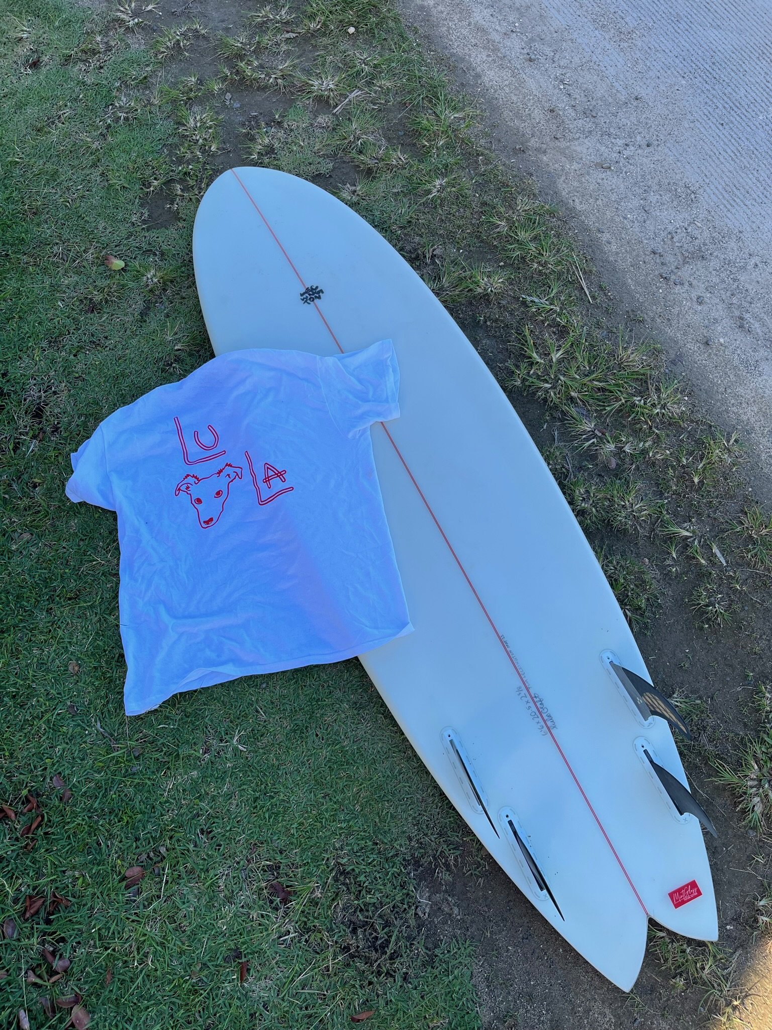 A white stand-up paddleboard lying on the grass next to a dirt path, with a plastic bag resting on the board.