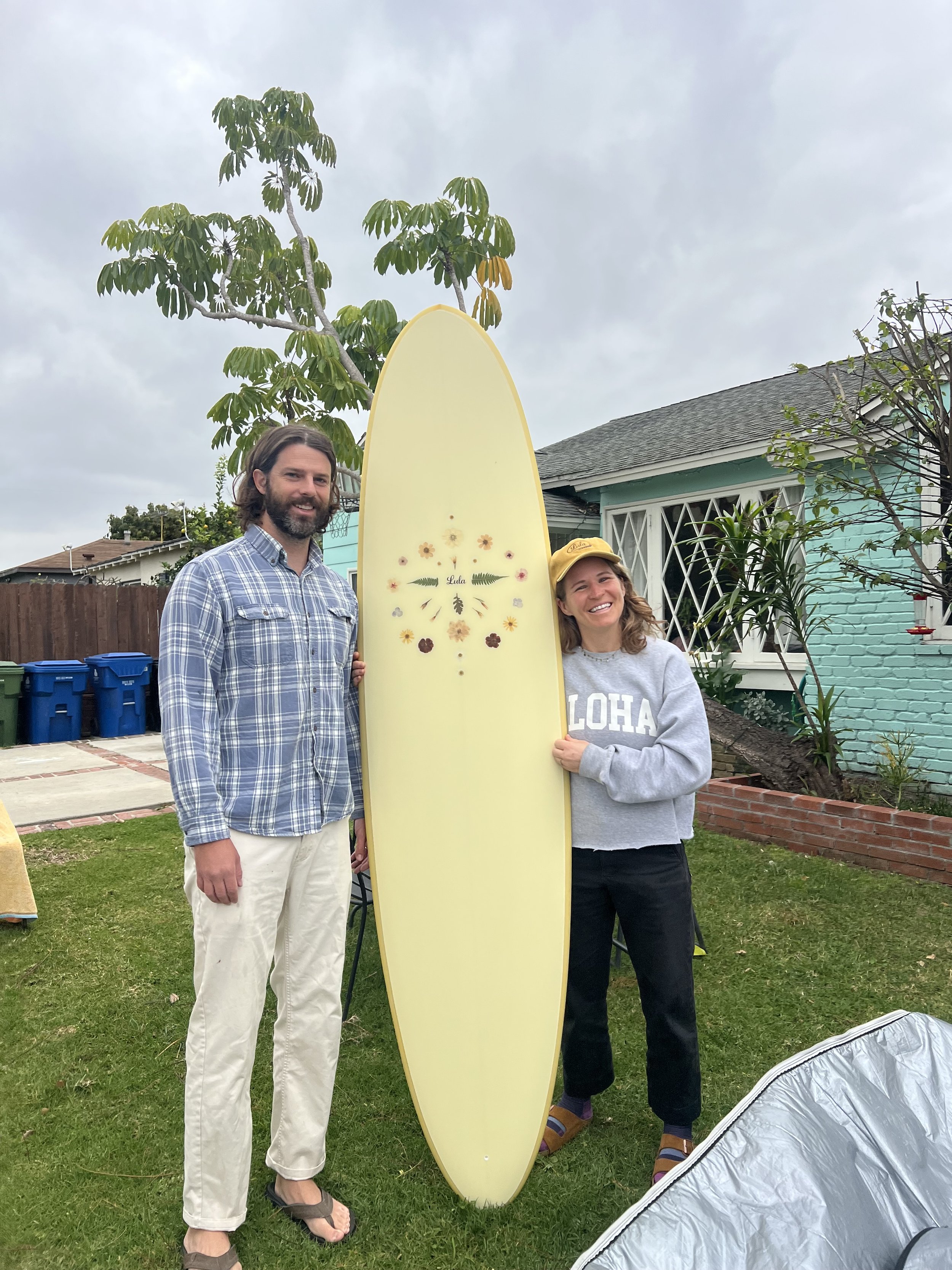 Two people, a man and a woman, standing outdoors next to a yellow surfboard with floral decorations, in front of a blue house with a gray roof and a yard with trees and plants, on a cloudy day.