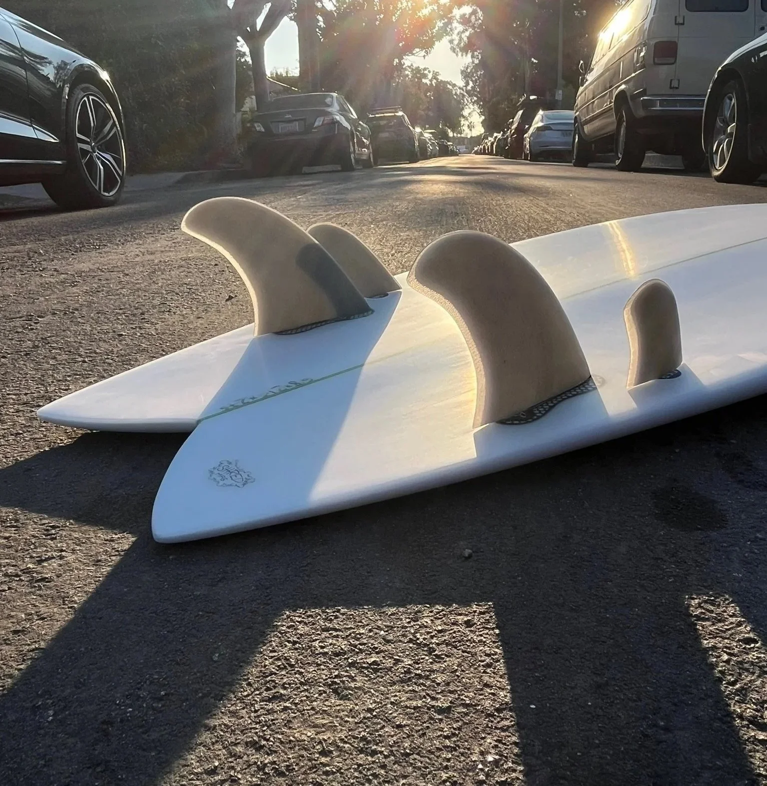 A white surfboard with three beige fins lying on a paved road, with parked cars on both sides and trees in the background, sunlight shining through the trees.