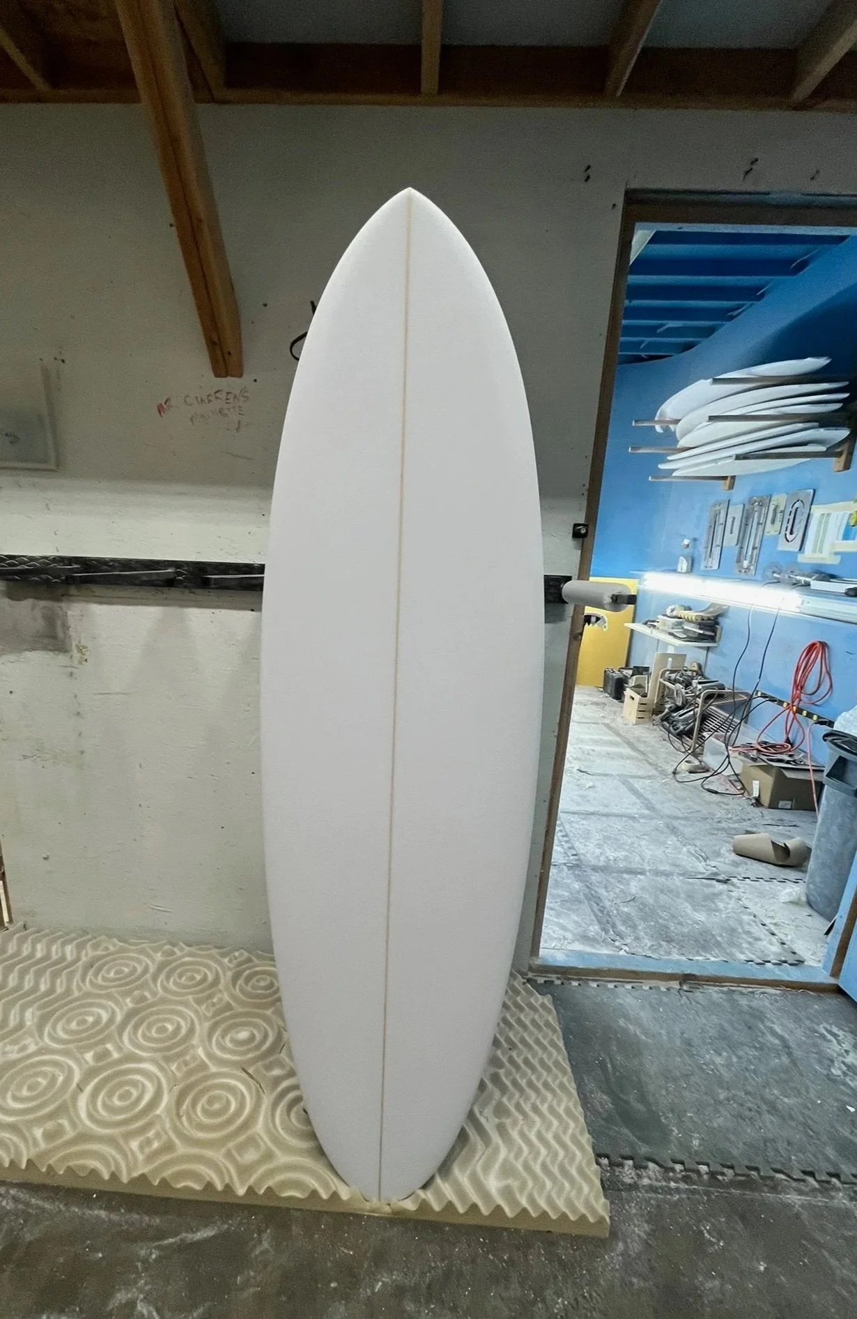 Unfinished white surfboard standing upright in a workshop area with tools and skateboard decks on the walls.