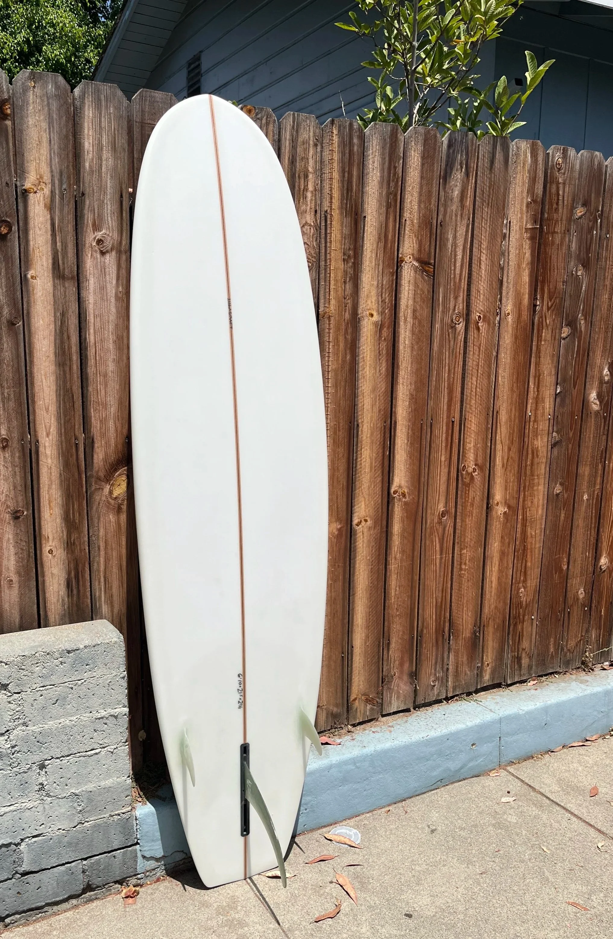 A white surfboard with a single brown stripe leaning against a wooden privacy fence. The skateboard is positioned on a concrete sidewalk, with some dried leaves scattered on the ground. The background includes part of a house with horizontal siding and a garden with a plant.