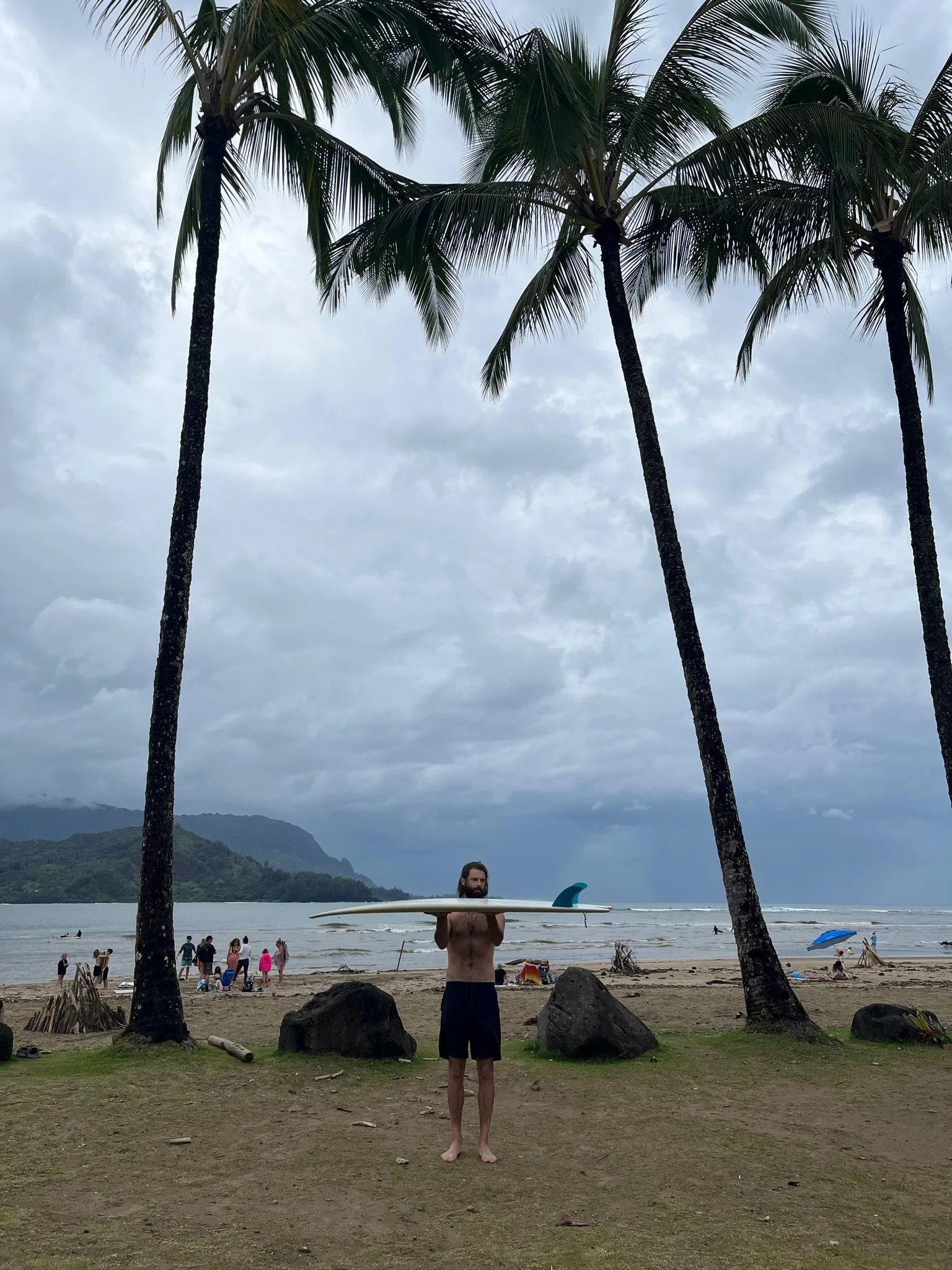 A person standing on a sandy beach holding a surfboard, with tall palm trees and cloudy sky in the background. The beach has several people, some with umbrellas and others near the water, and a distant mountain range is visible across the water.