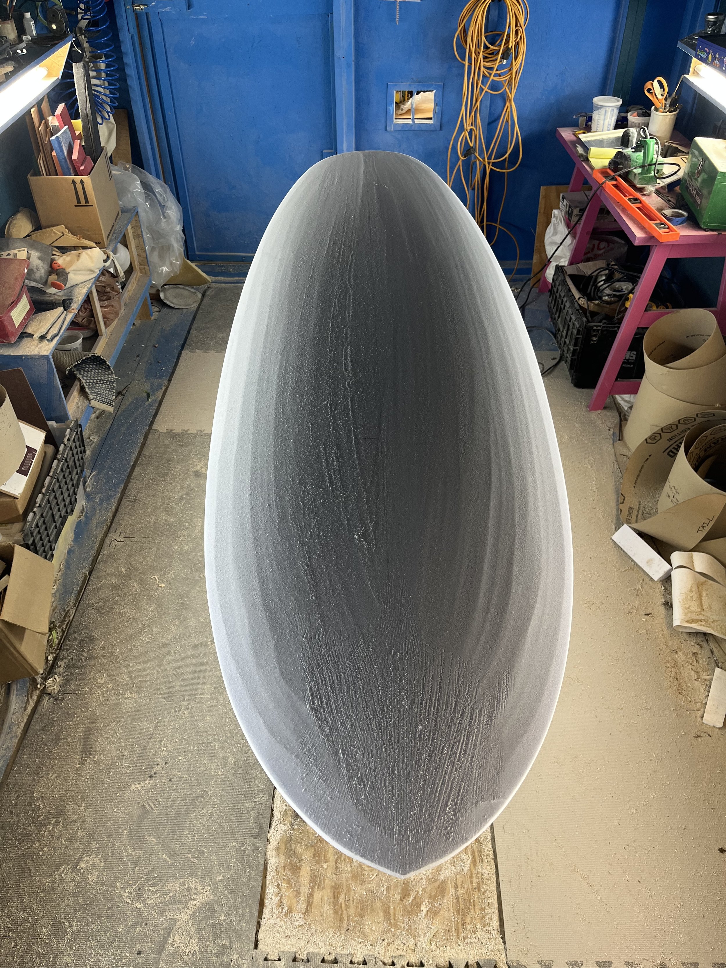 A workshop scene showing the top view of a boat hull in progress, with workbenches and tools surrounding it.