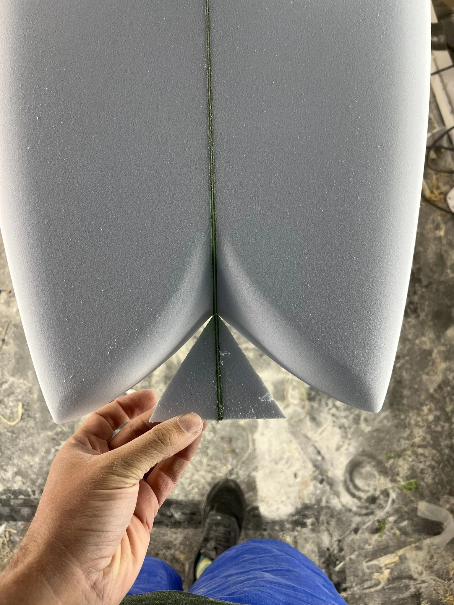 A close-up view of the bottom of a surfboard being held by a hand. The surfboard has a gray, textured surface with a faint green line running down the center.