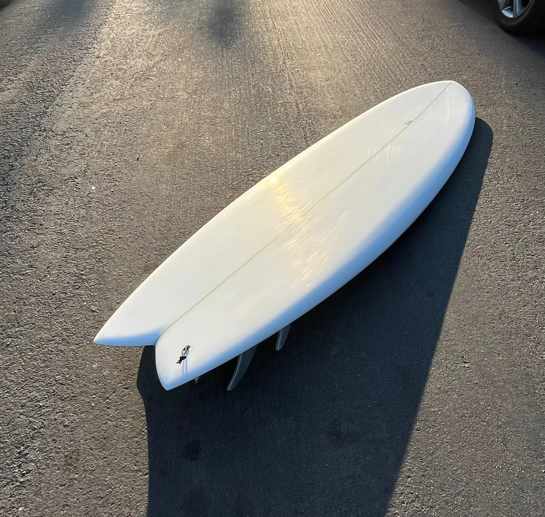 A white surfboard lying on the asphalt ground with shadows cast from the sunlight, near a parked car.