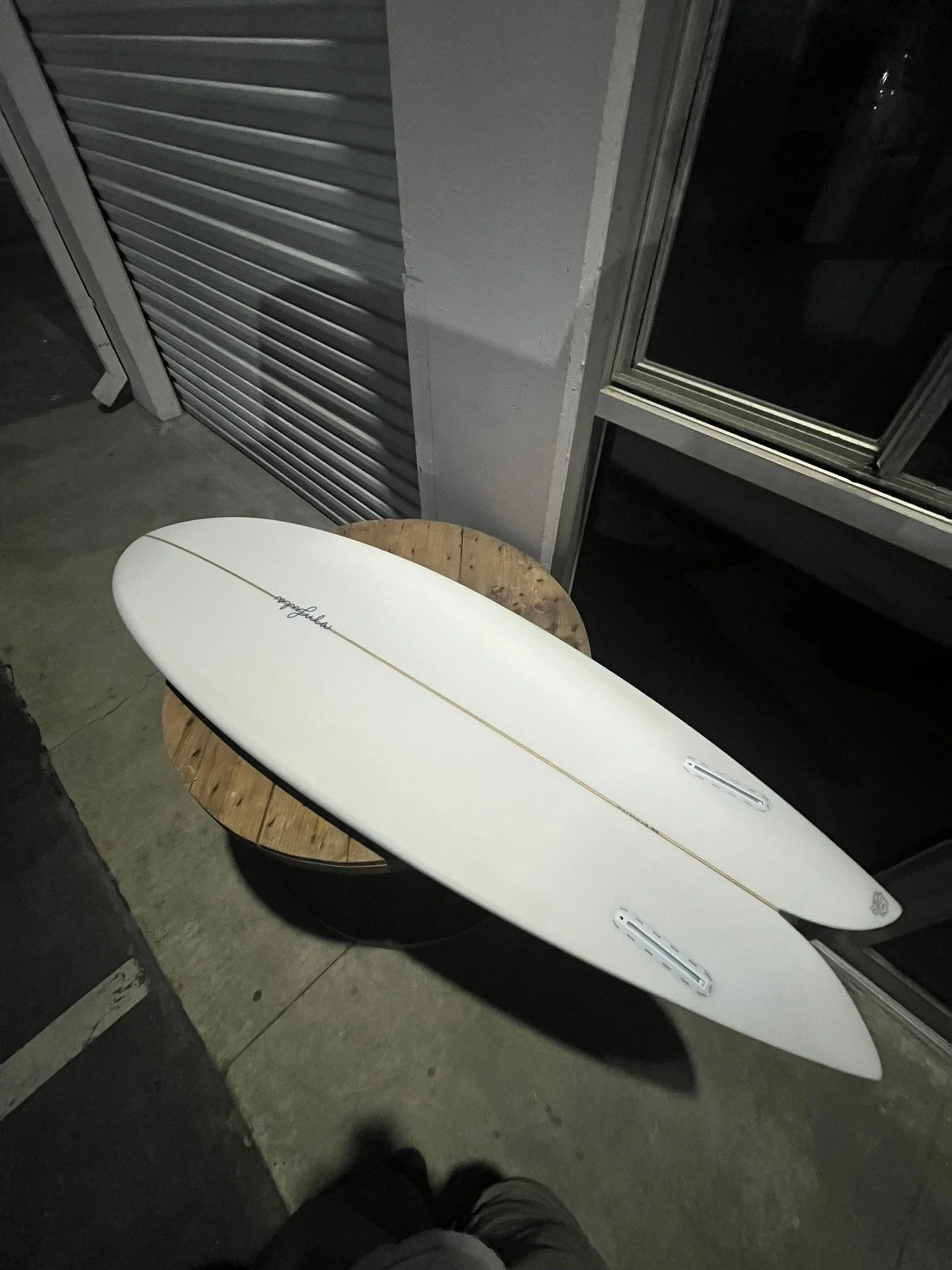 A white surfboard with a black fin and a logo, leaning against a round wooden table, in a dimly lit indoor space with metal window shutters.