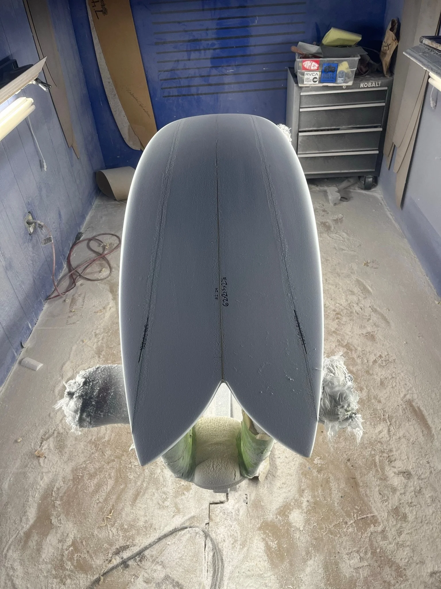 A surfboard painted light gray with black lines, lying upside down on a chair in a workshop. The workshop has a blue wall, a black tool chest, and various tools and supplies around.