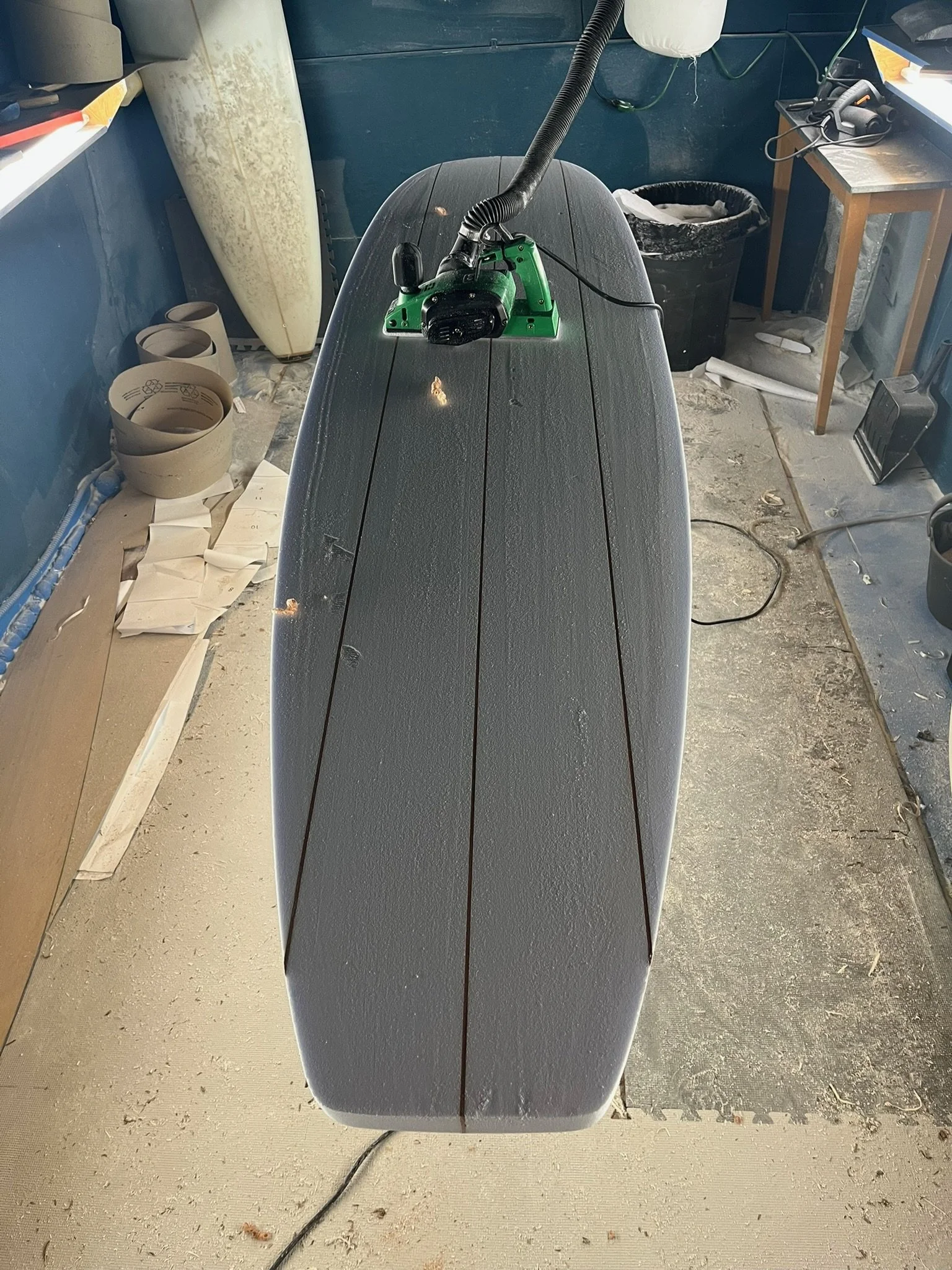 A surfboard with a mounted vacuum hose and ruger attached, situated on a worktable in a workshop. The workshop has various tools, a trash can, and a desk in the background.