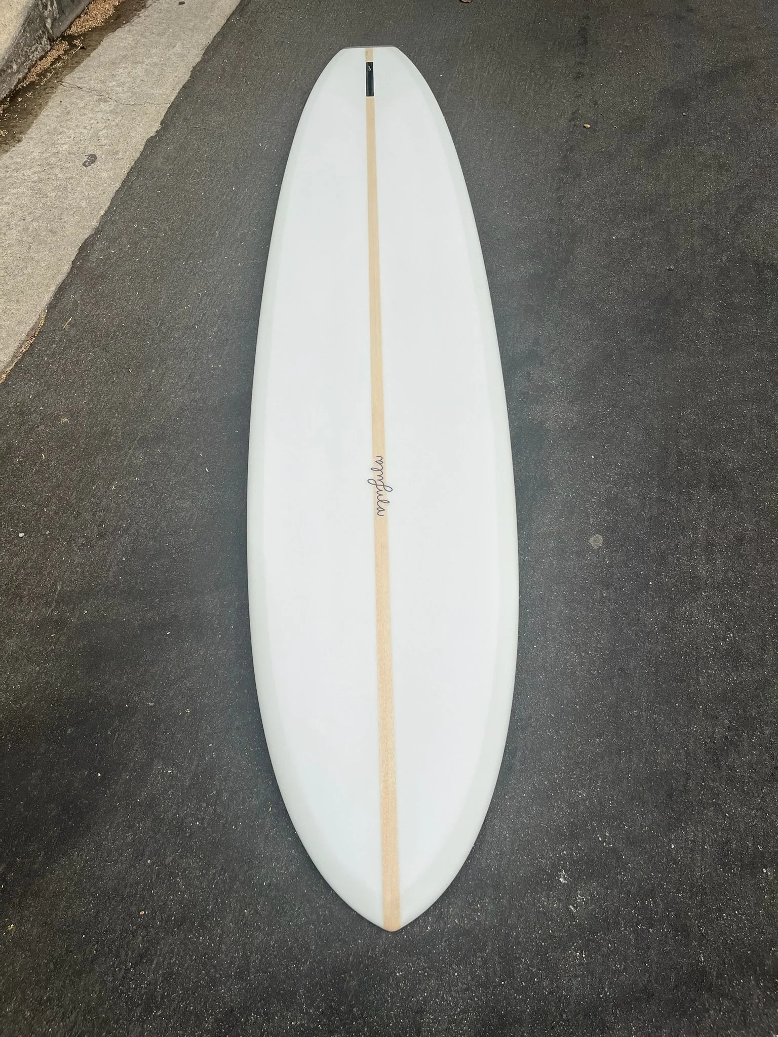 A white surfboard with a natural wood stripe running down the center, lying on a dark asphalt surface.