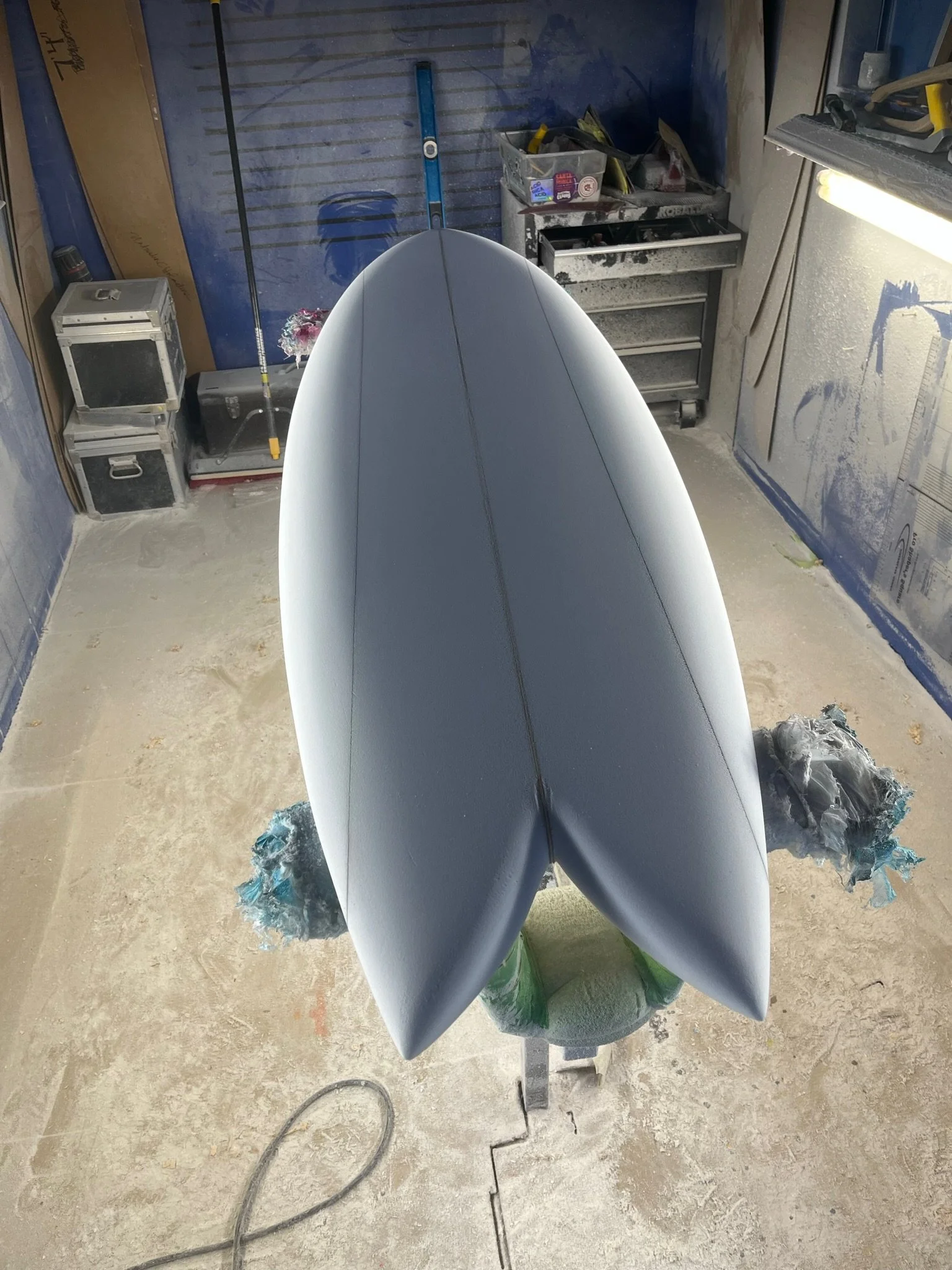 The image shows an unfinished surfboard in a workshop, positioned vertically on a stand, with tools, storage boxes, and supplies visible in the background.