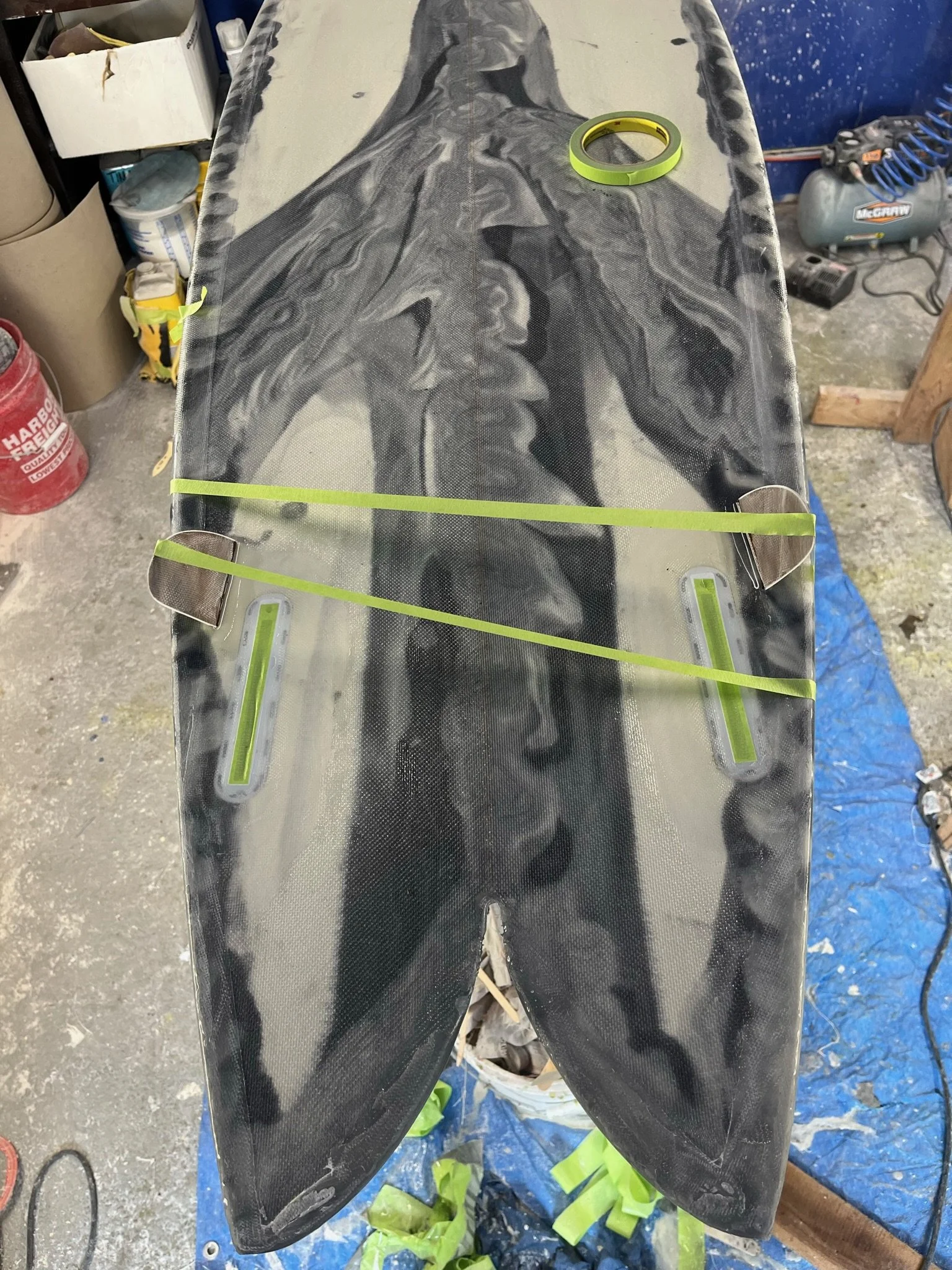 A custom painted surfboard with tape around the edges for fin alignment, placed on a workbench in a garage or workshop.