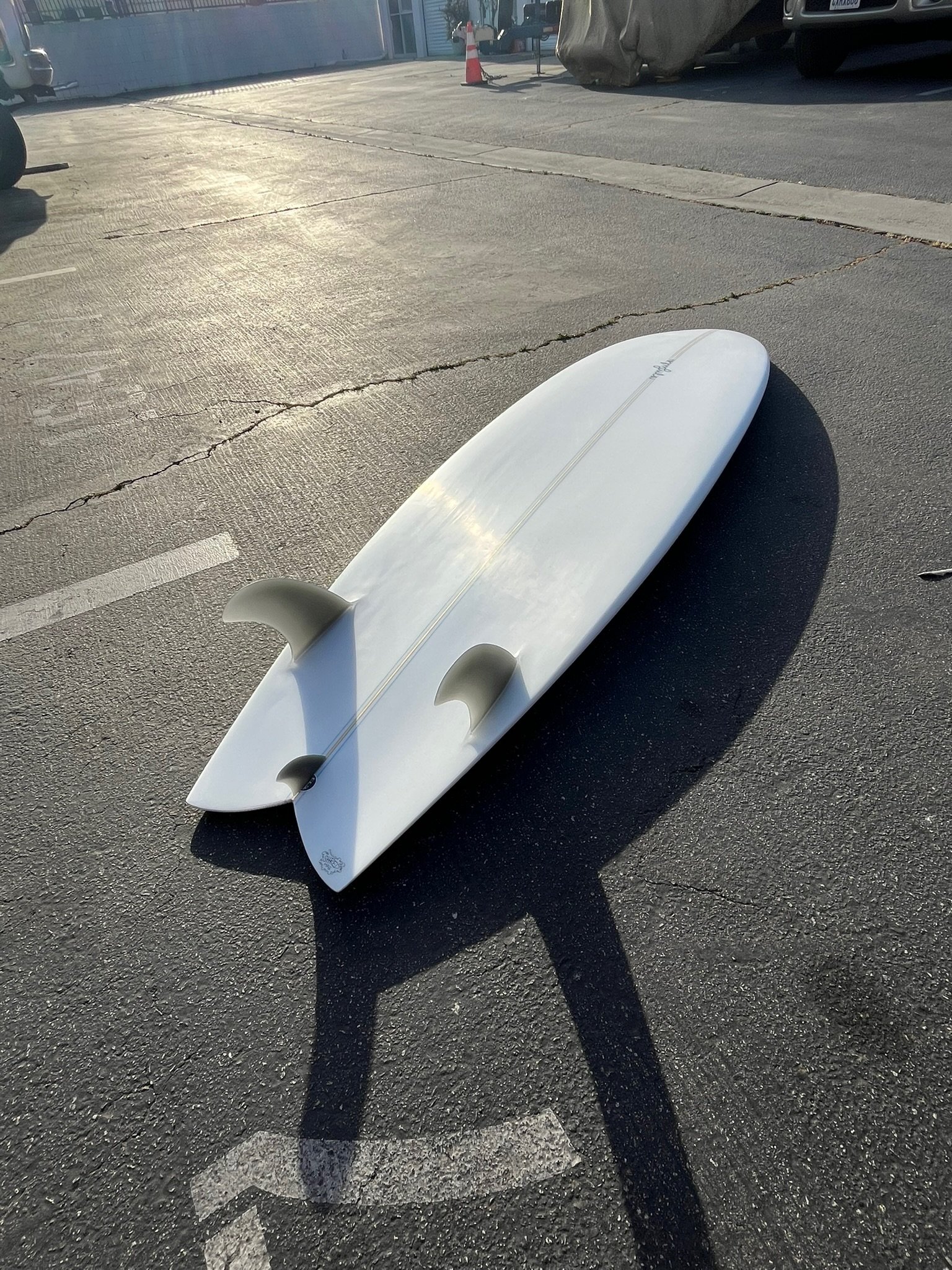 A white surfboard lying on a parking lot pavement, casting a shadow in the sunlight. A traffic cone and some vehicles are visible in the background.