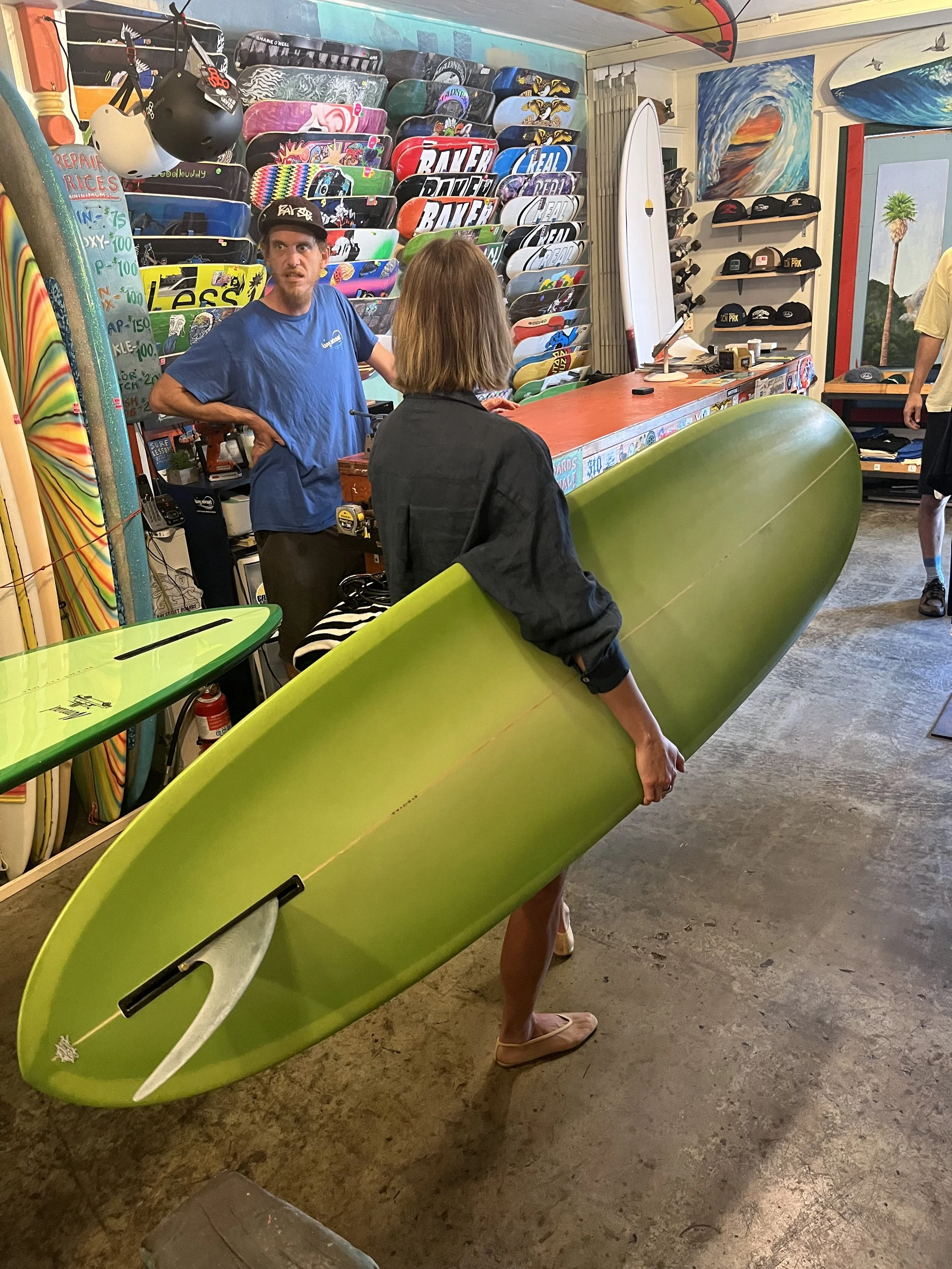 A person holding a bright green surfboard inside a surf shop. The surfboard has a white area on the nose and is being held by a woman wearing flip-flops and a black jacket. The shop has racks of skateboards, caps, and surf accessories, with a colorfu