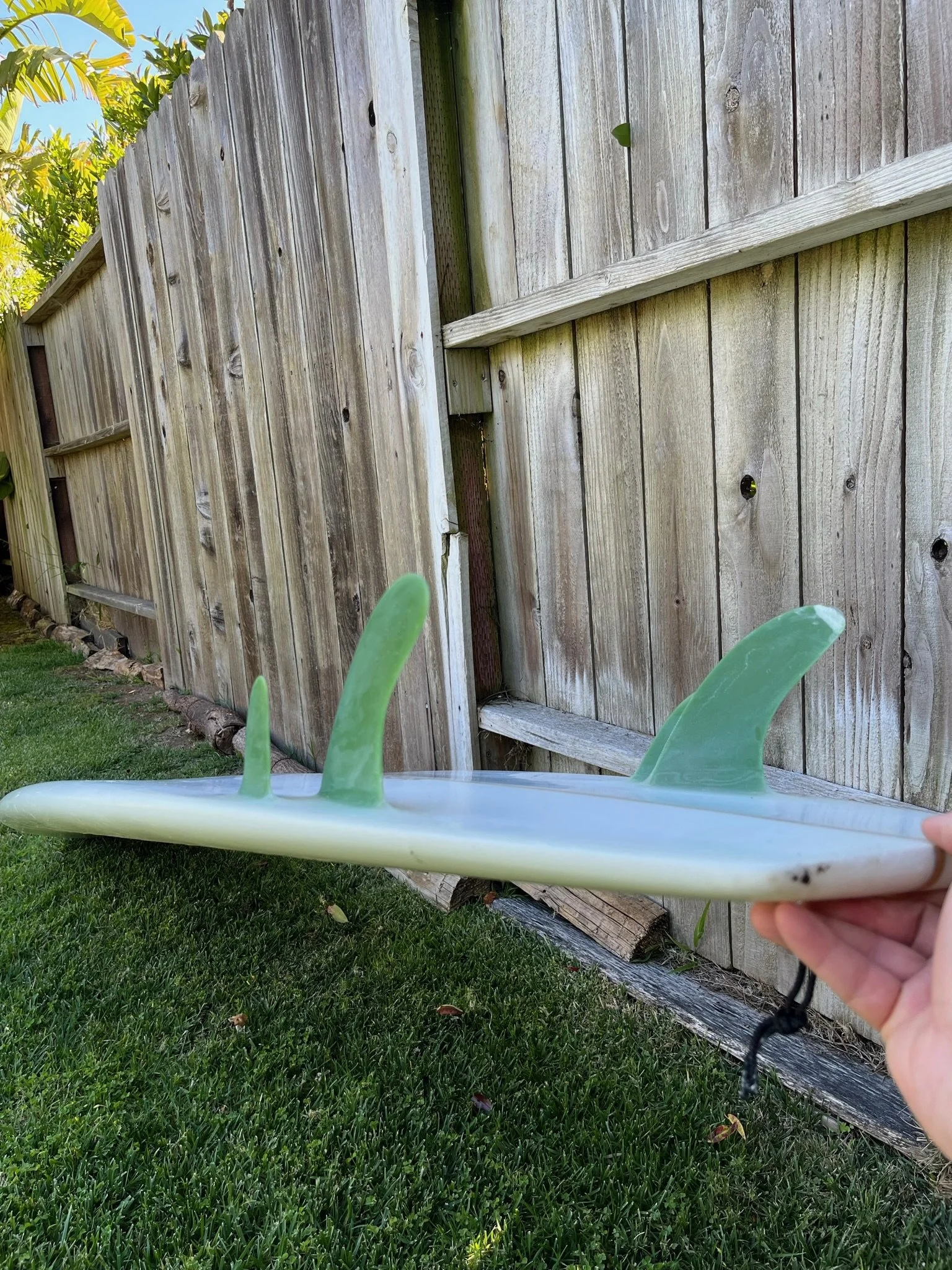 A person holding a surfboard upside down with green quad fins in front of a weathered wooden fence and green grass.