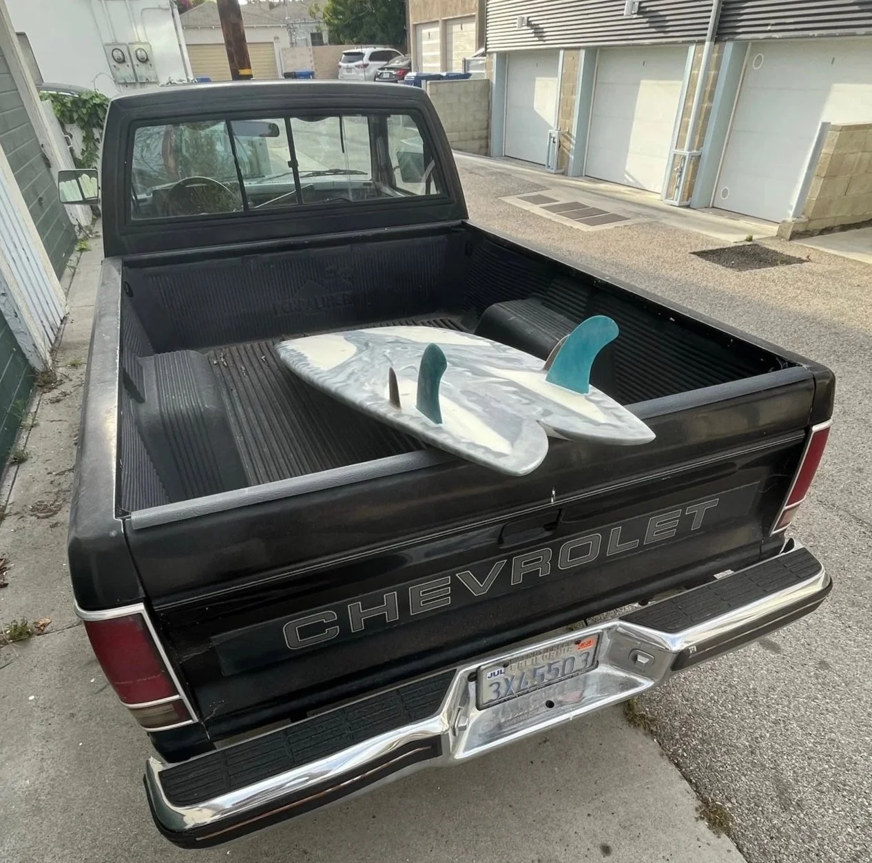 Black Chevrolet pickup truck with two surfboards in the bed, parked in a driveway alongside a sidewalk and residential buildings.