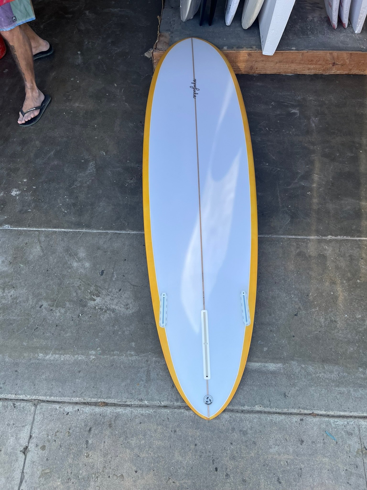 A surfboard with a white top and yellow edge lying on the ground. Visible part of a person's leg and foot in flip-flops on the left, and a set of white surfboards on a rack in the background.