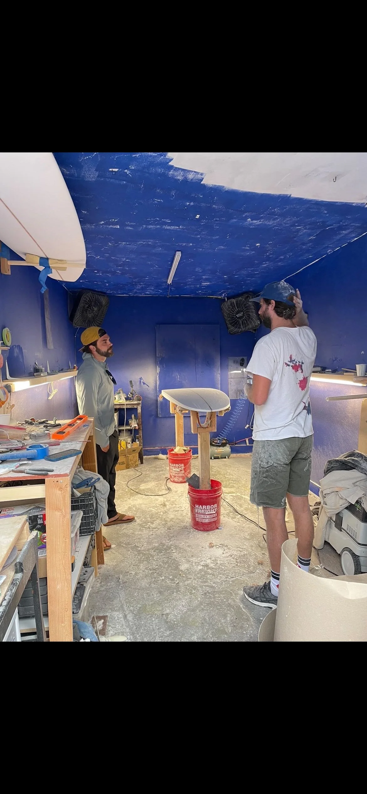 Two men in a workshop with blue walls and ceiling, painted partially with dark blue paint. One man wears a white t-shirt and shorts, the other wears a light-colored jacket and a yellow cap. The workshop has shelves with tools, paint buckets, and othe