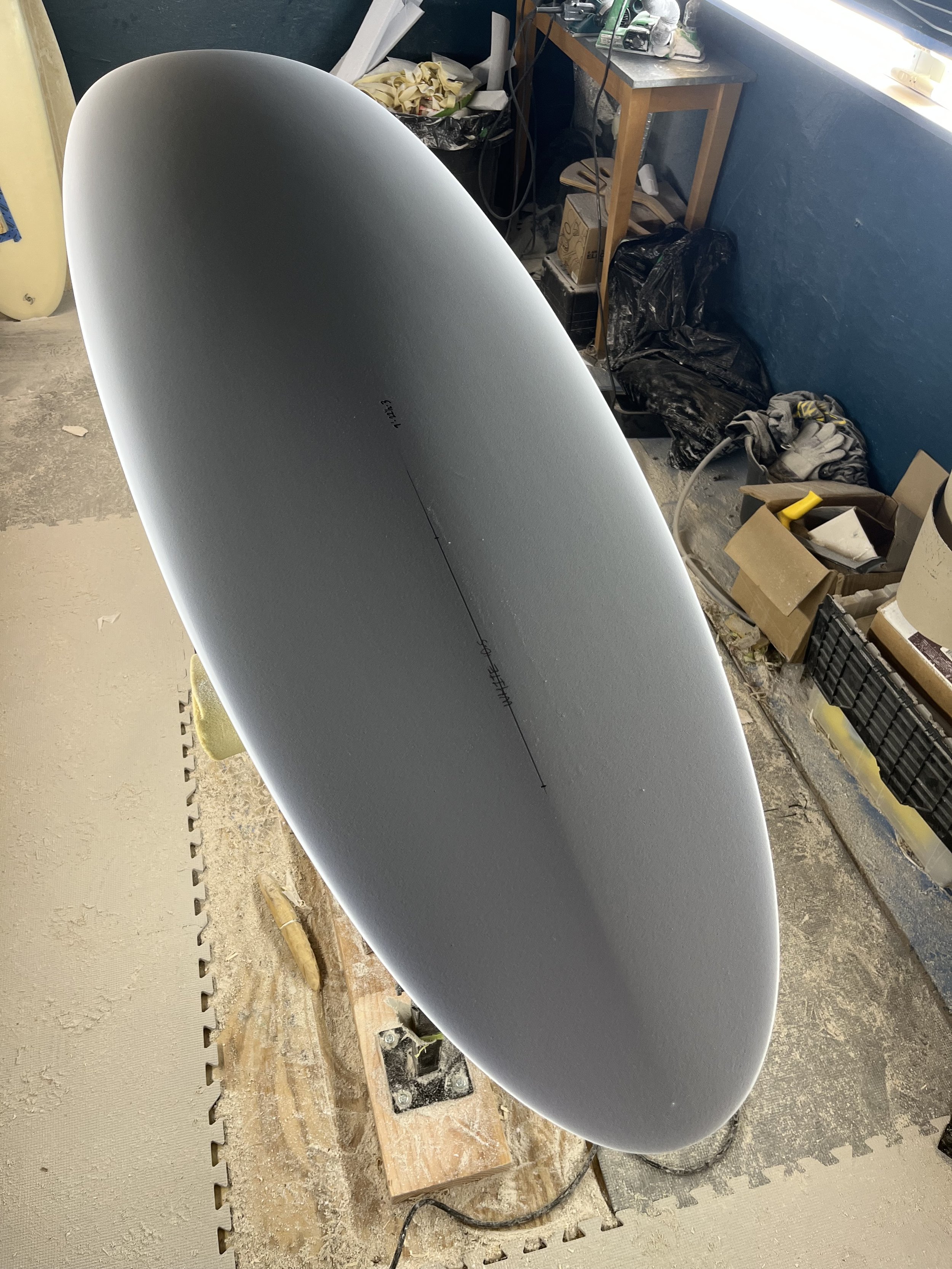 A surfboard is propped against a workbench in a workshop. The workshop has various tools, supplies, and clutter, including gloves, boxes, and plastic bags. The floor appears rough and dusty.