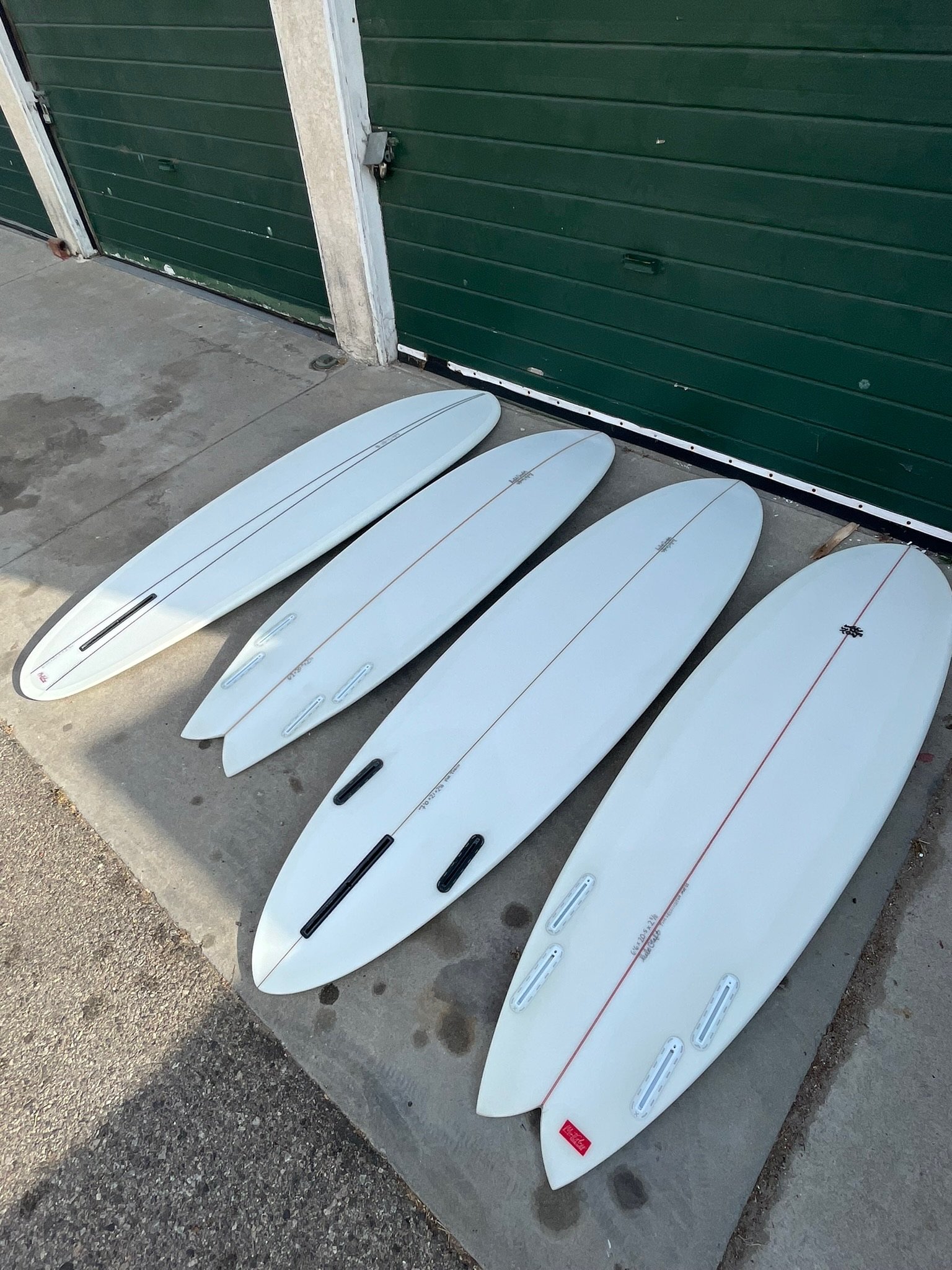 Six white surfboards with black, orange, and white fin setups lying on the ground in front of a green garage door.