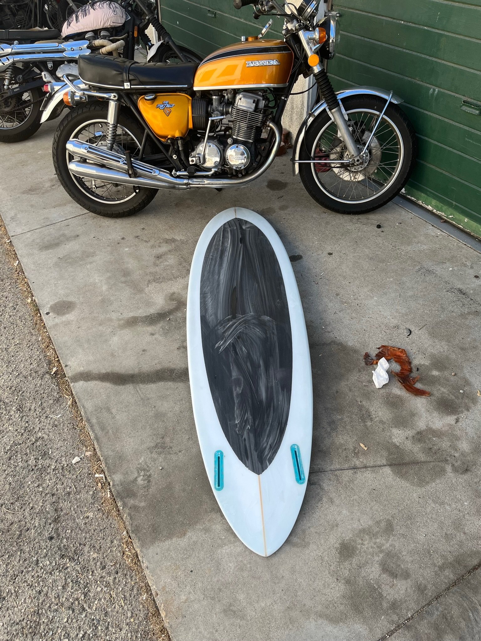 A vintage orange and black Honda motorcycle parked next to a green wall, with a paddleboard lying on the ground in front.