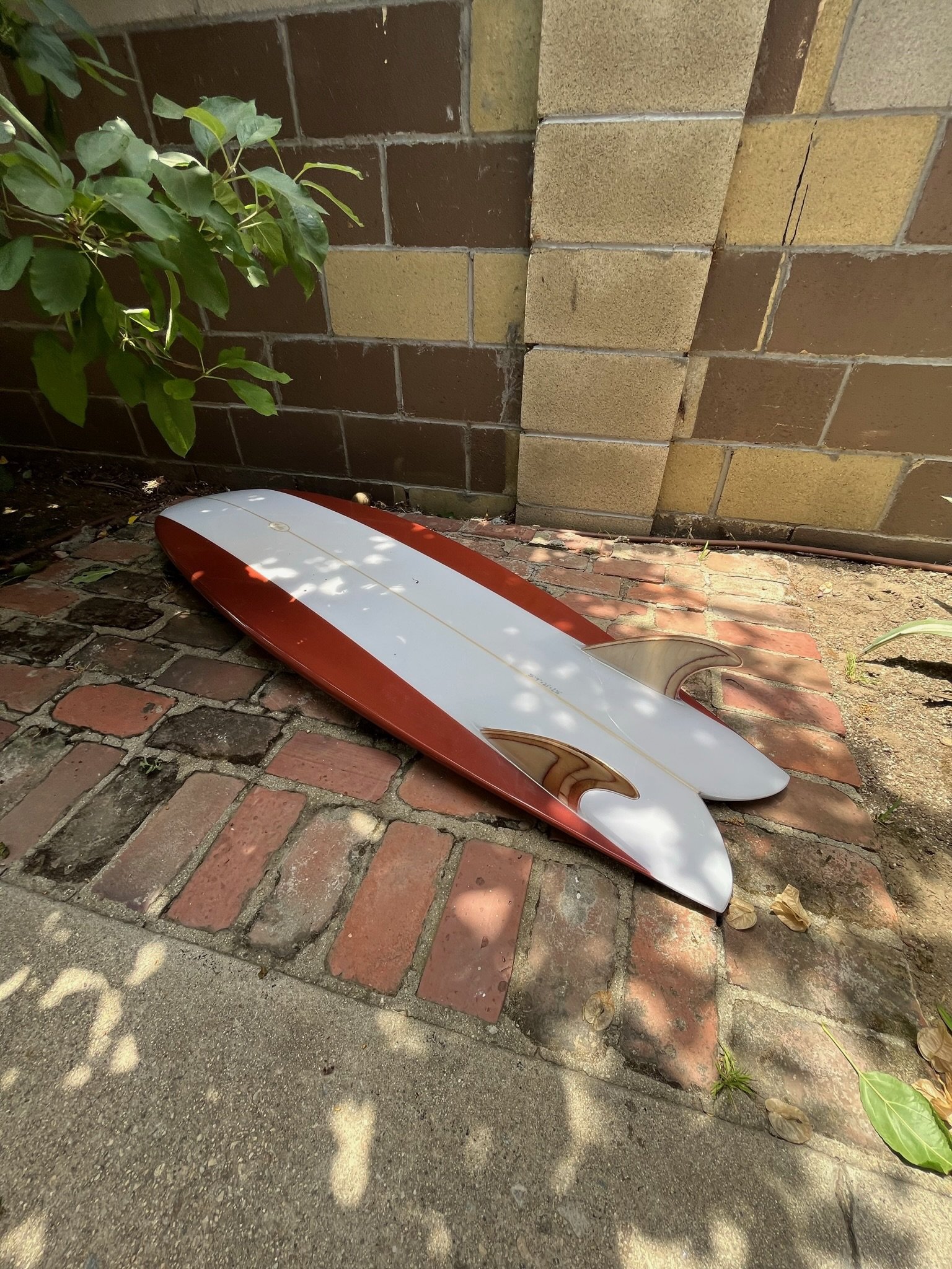 A fish style short twin fin surfboard with red and white colors leaning against a brick and tile wall. The ground is covered with brick and asphalt with some shadows and leaves.