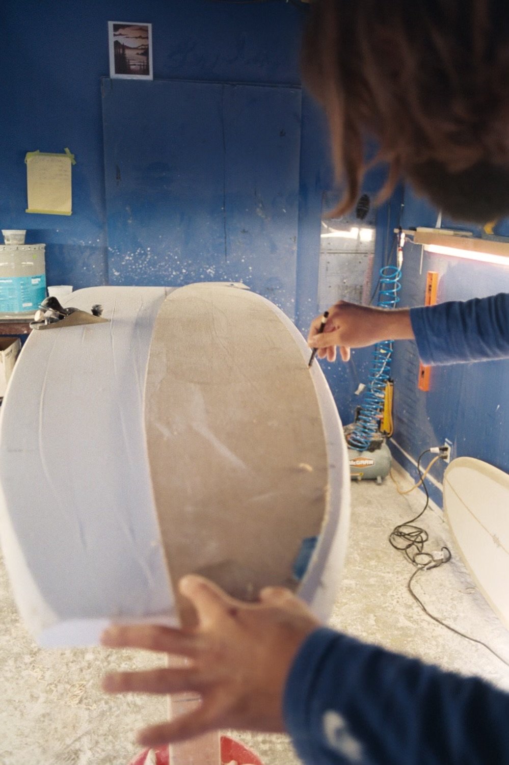 Person shaping a foam surfboard blank in a workshop with blue walls, tools, and supplies in the background.
