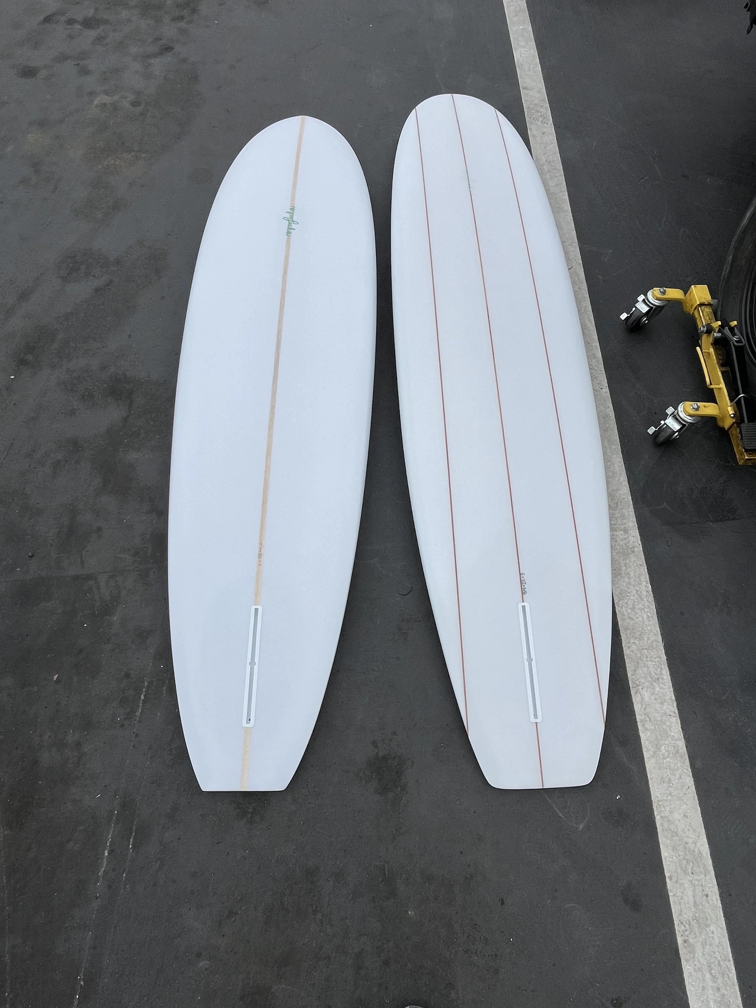 Two surfboards lying on a parking lot pavement, one with a solid white color and a thin stripe along the center, the other also white with red lines running lengthwise.