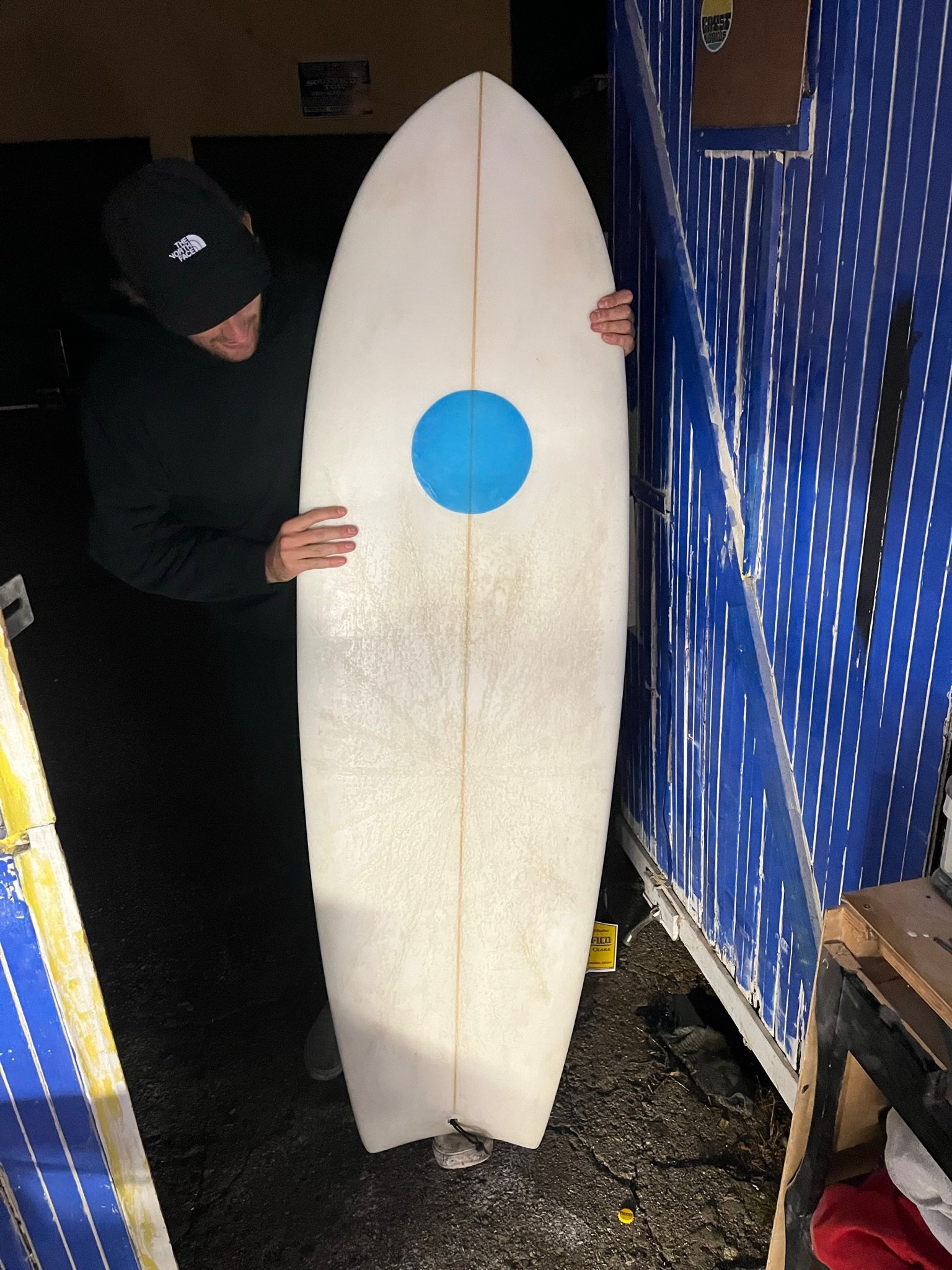 A man in a black North Face hoodie and a black beanie hat holding a white surfboard with a blue circle in the center, standing in a storage area with blue and brown containers.