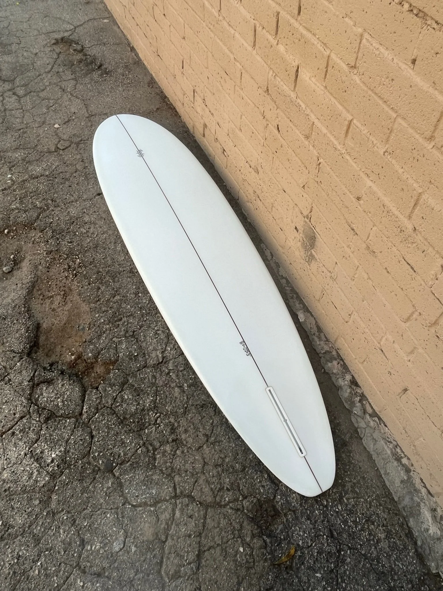 A white surfboard with a black string runs down the center, leaning against a unfinished brick wall on a cracked asphalt surface.