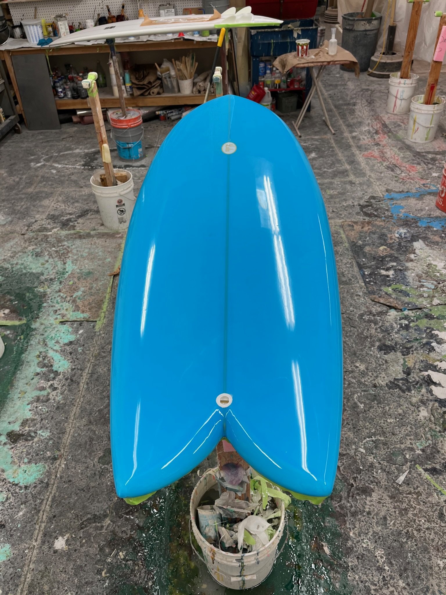 A blue surfboard mounted on a stand above a paint bucket filled with paper and paint supplies in a workshop.