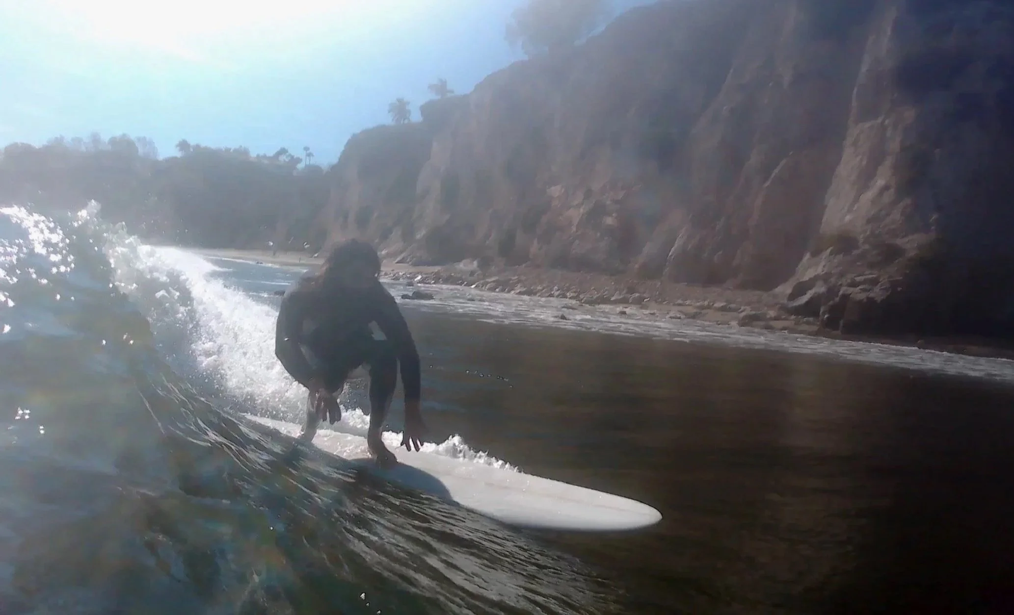 A person surfing on a small wave near a rocky coastline with cliffs and trees in the background.