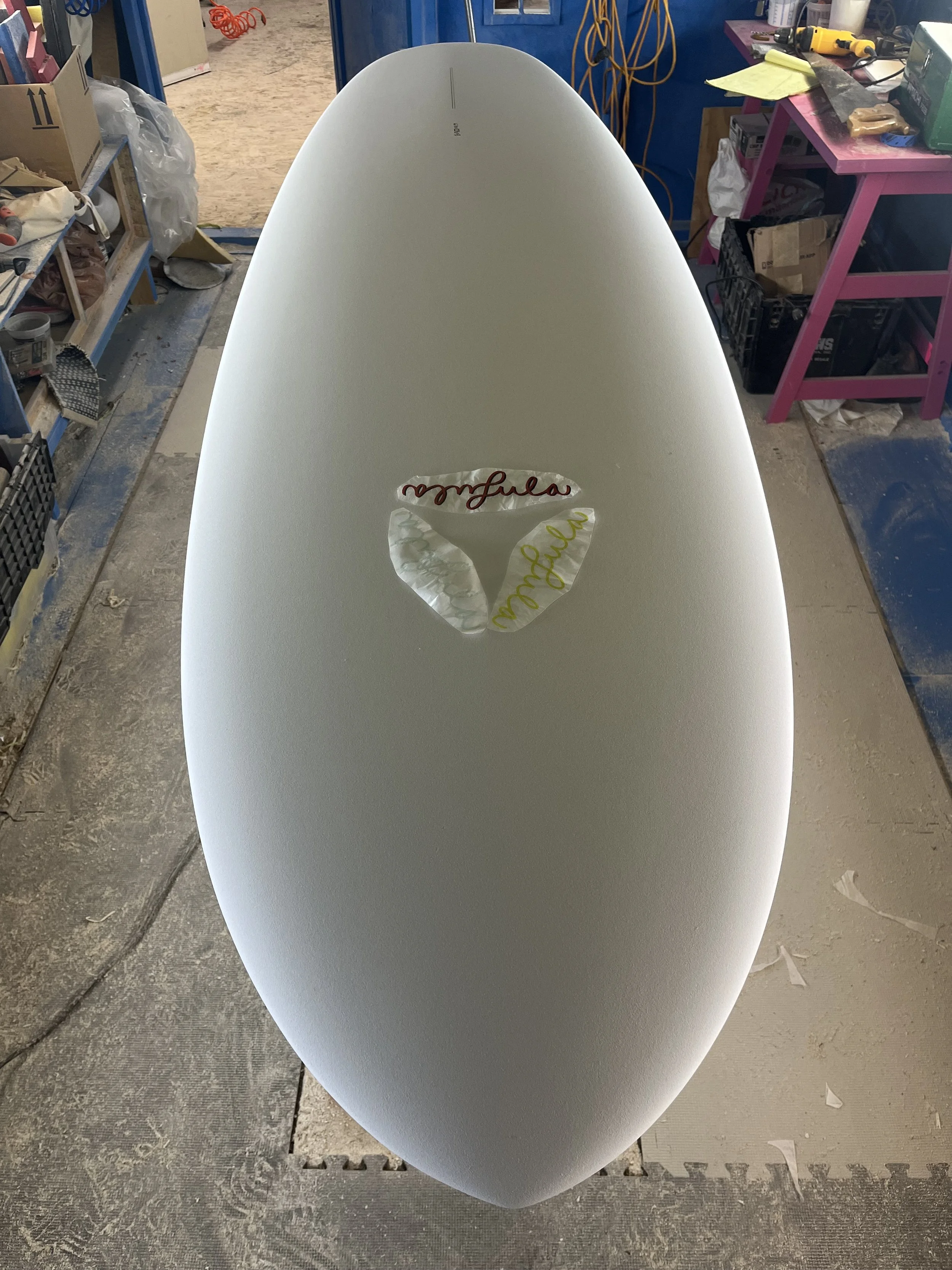 A surfboard in a workshop, with a sticker on top that reads 'mfluence' in red and yellow writing.
