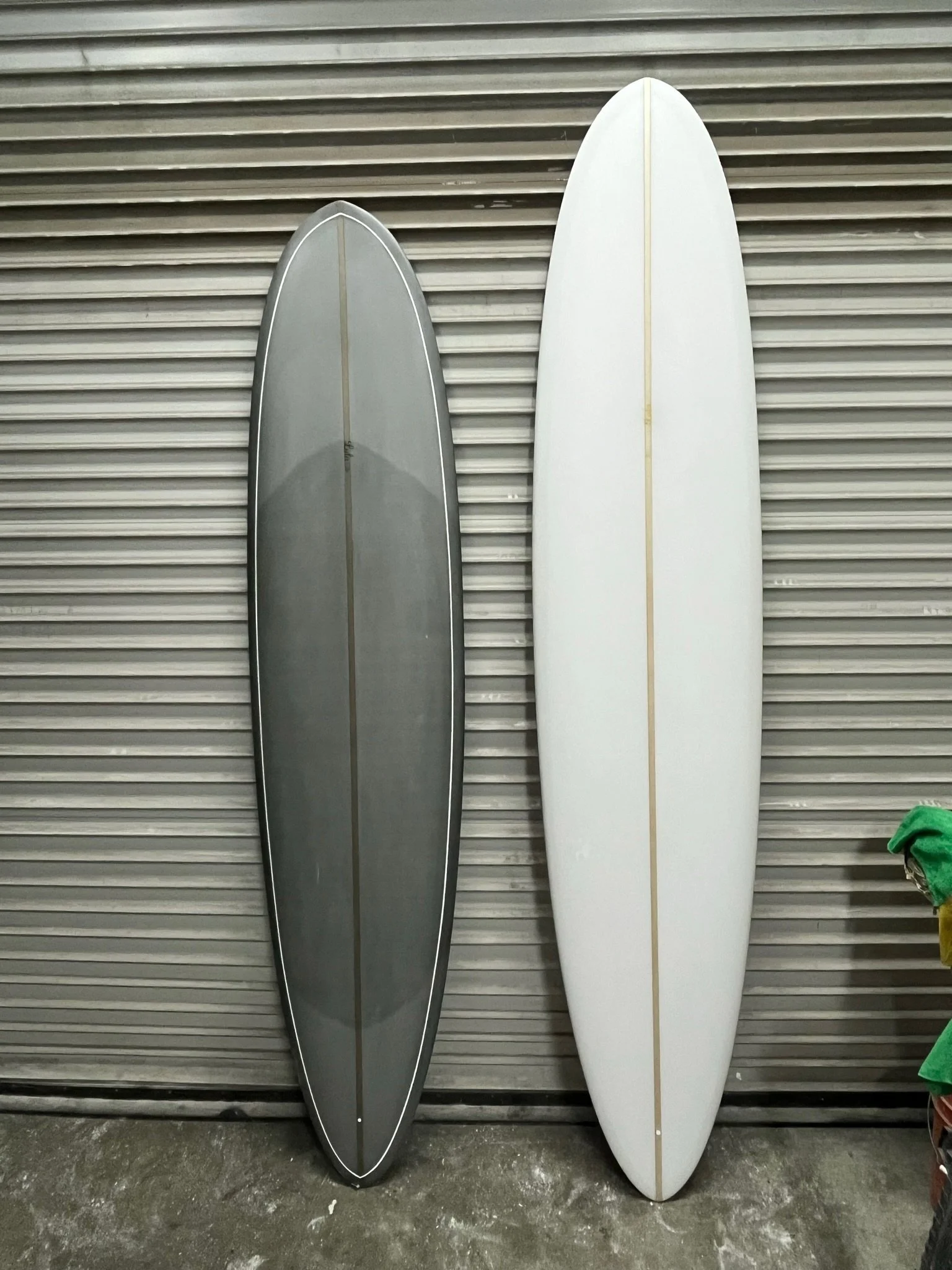 Two surfboards leaning against a metallic, corrugated wall, one gray and one white, with a green cloth or towel partially visible on the right side.