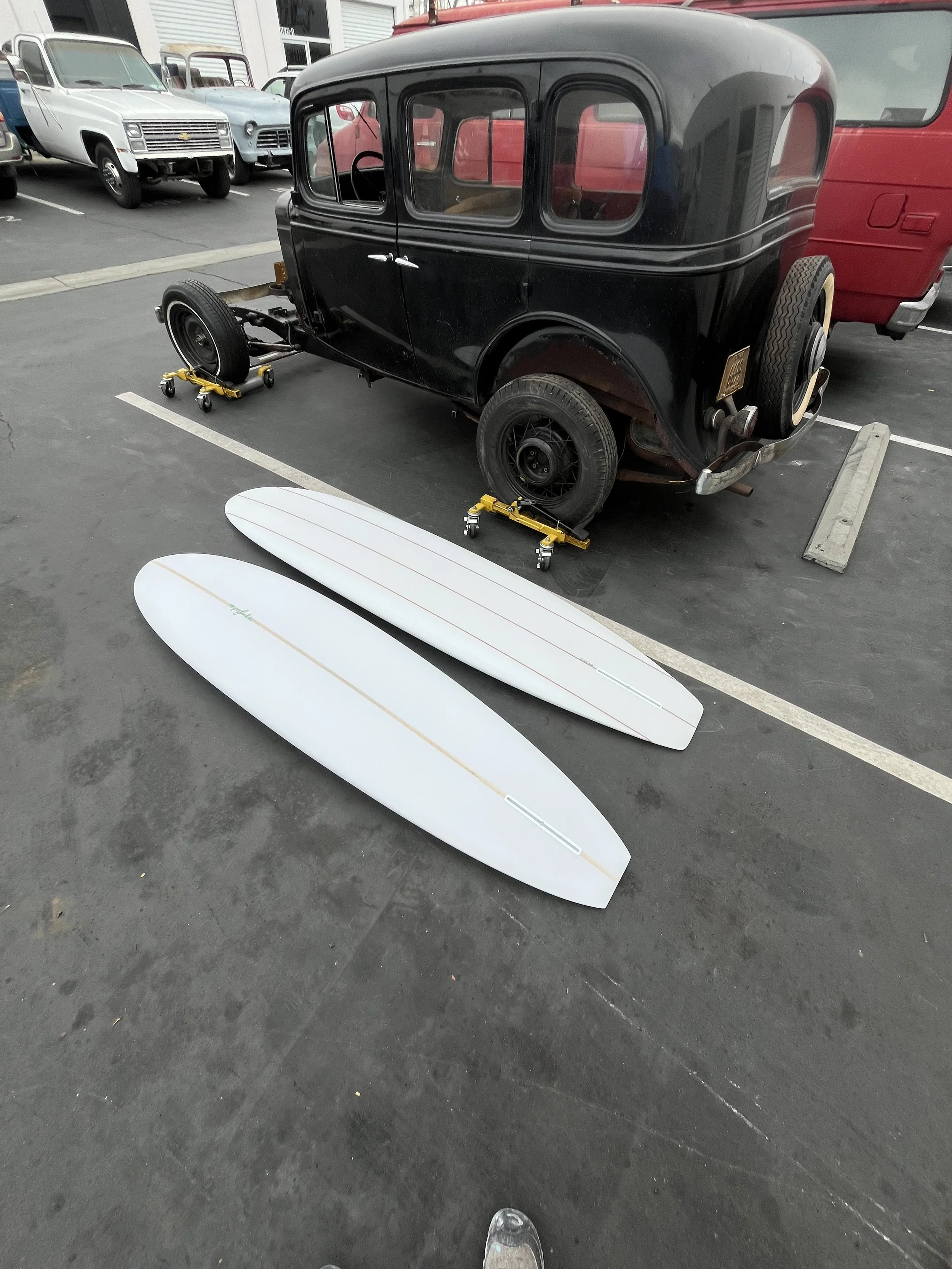 A vintage black car with a rounded body and classic design, partially disassembled on a parking lot, with surfboards on the ground nearby.