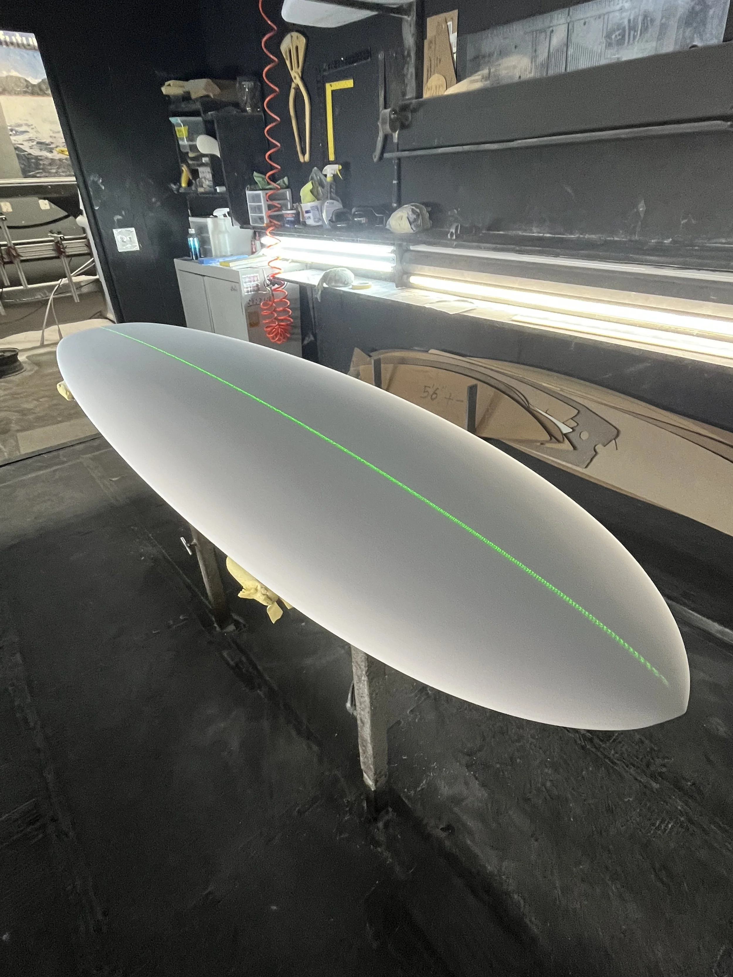 Workshop with various tools, fluorescent lights, and a surfboard with a green laser line, on a workbench.