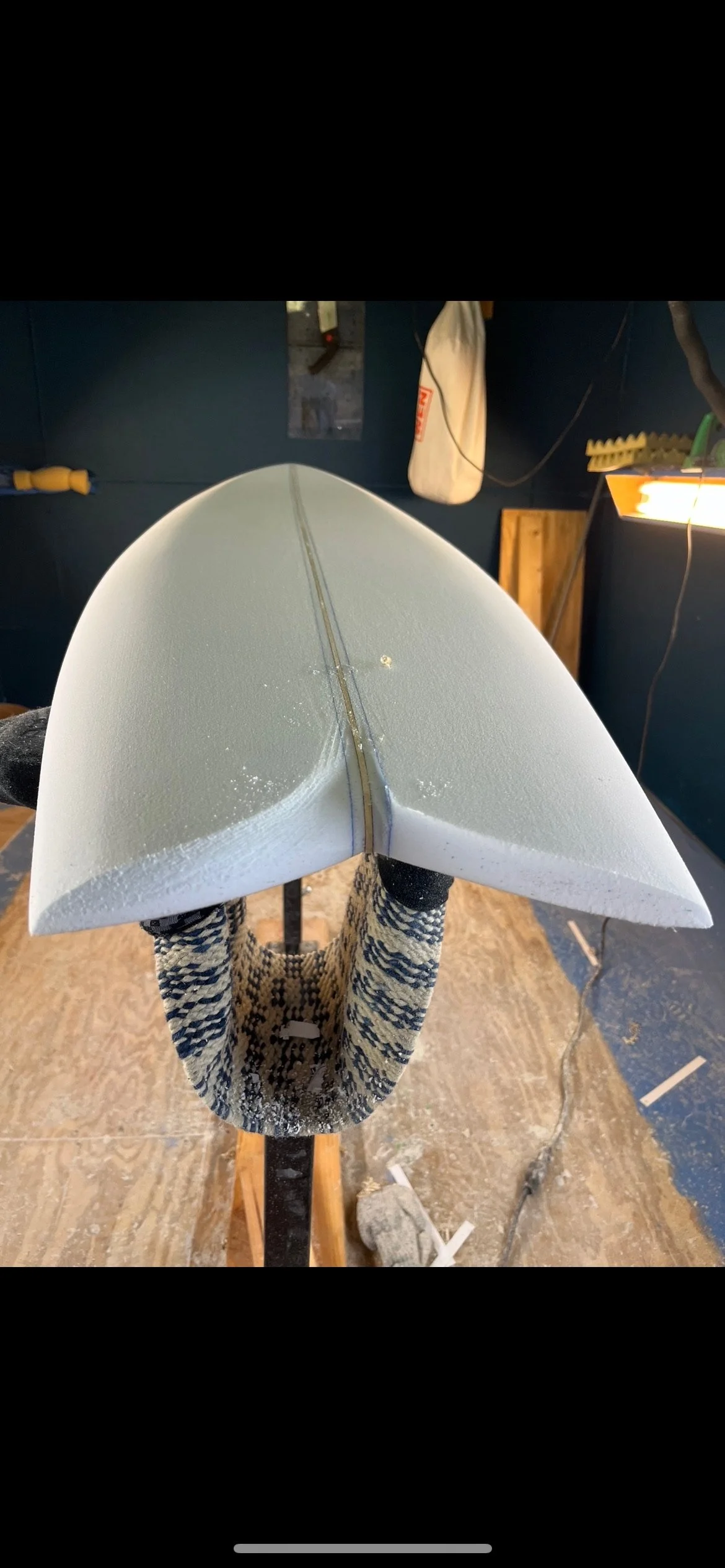 A surfboard in a workshop or garage, with a black and white patterned leash attached, and tools and equipment hanging on the wall behind it.