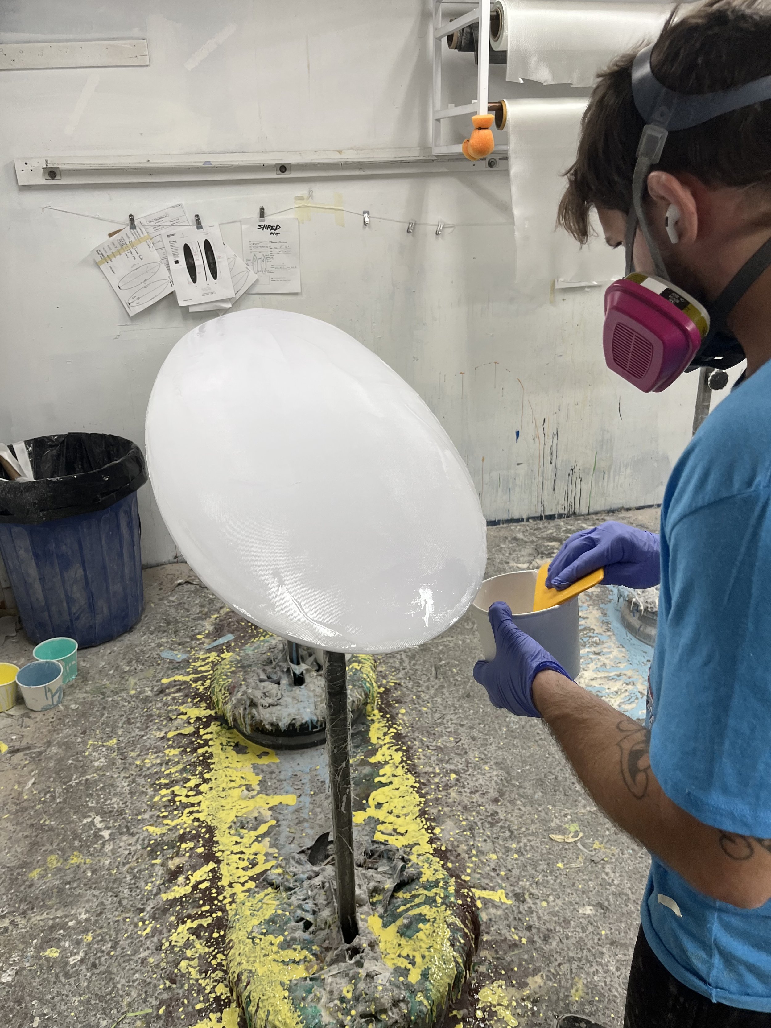 A person wearing a respirator mask and gloves creates a sculptural art piece, resembling a large eyeball, in an art studio with scattered paint and art supplies on the floor.