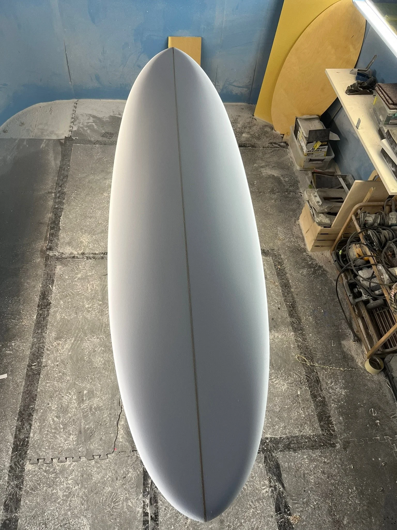 Top view of a white surfboard in a workshop setting, surrounded by tools and materials like sandpaper, power tools, and wooden boards.