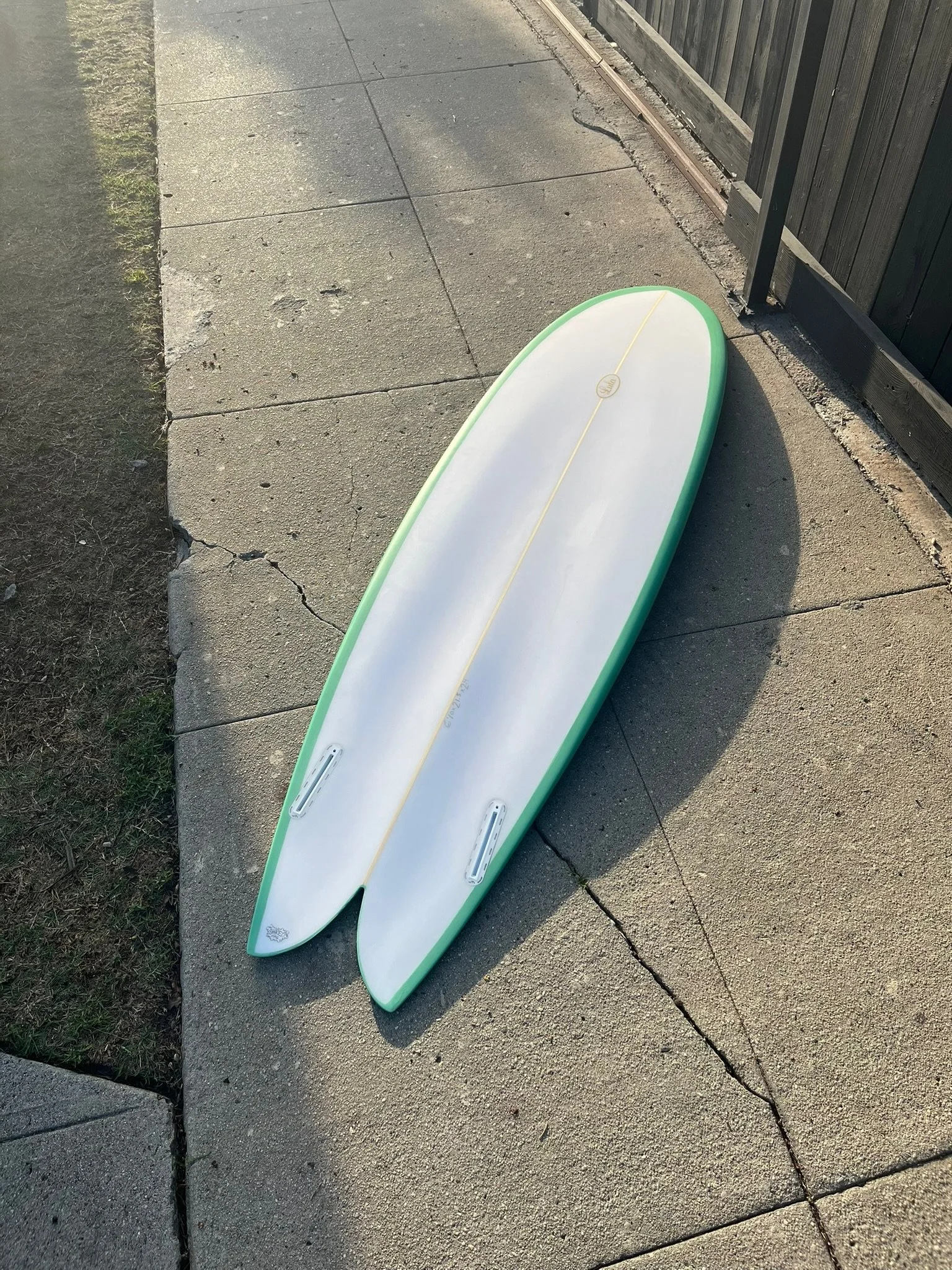Two surfboards lying on a concrete sidewalk next to a wooden fence and grass, one surfboard is white with a green edge, the other is not fully visible.
