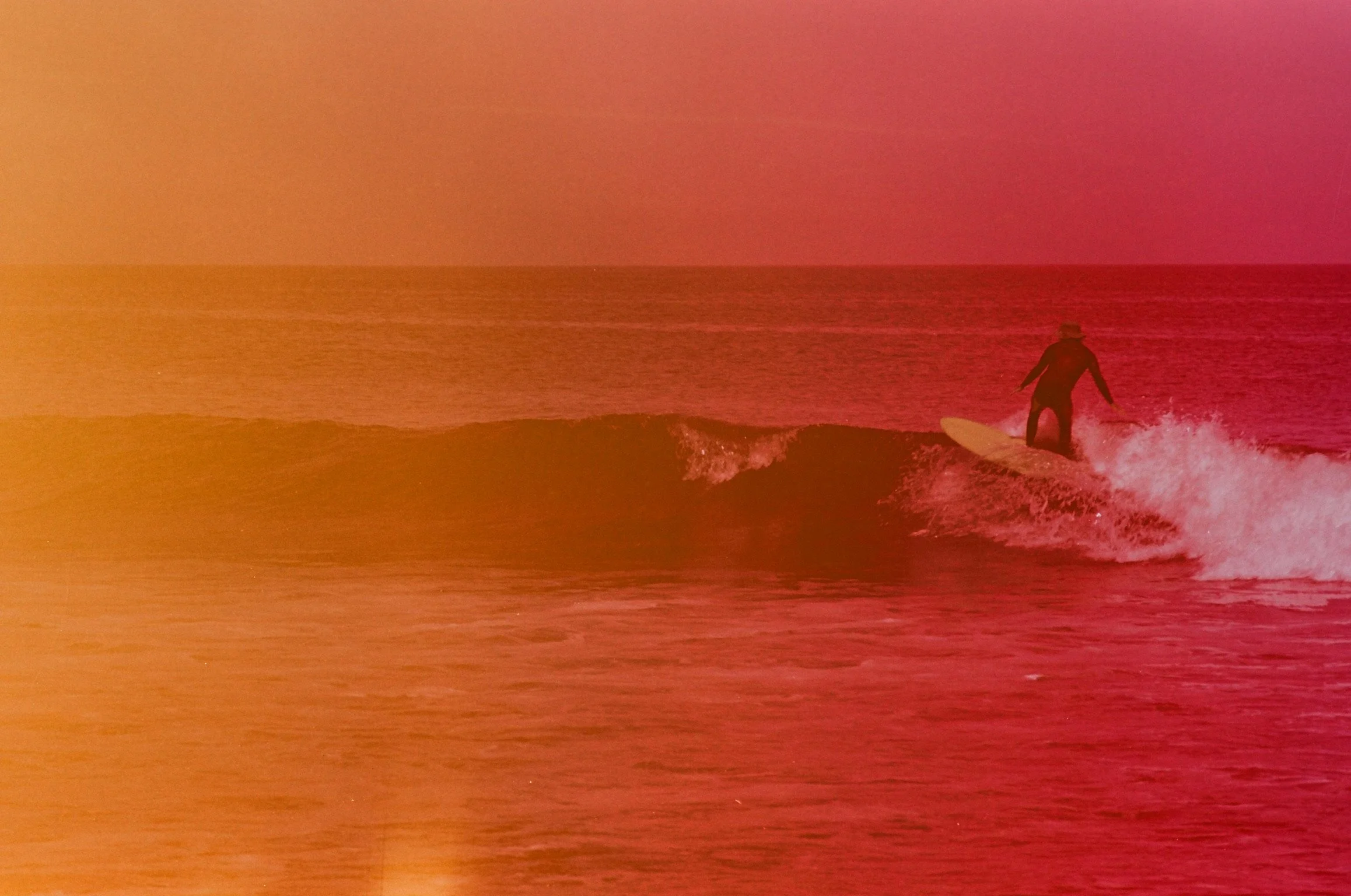 A person surfing on a wave at sunset or sunrise, with the sky illuminated in pink and orange hues.