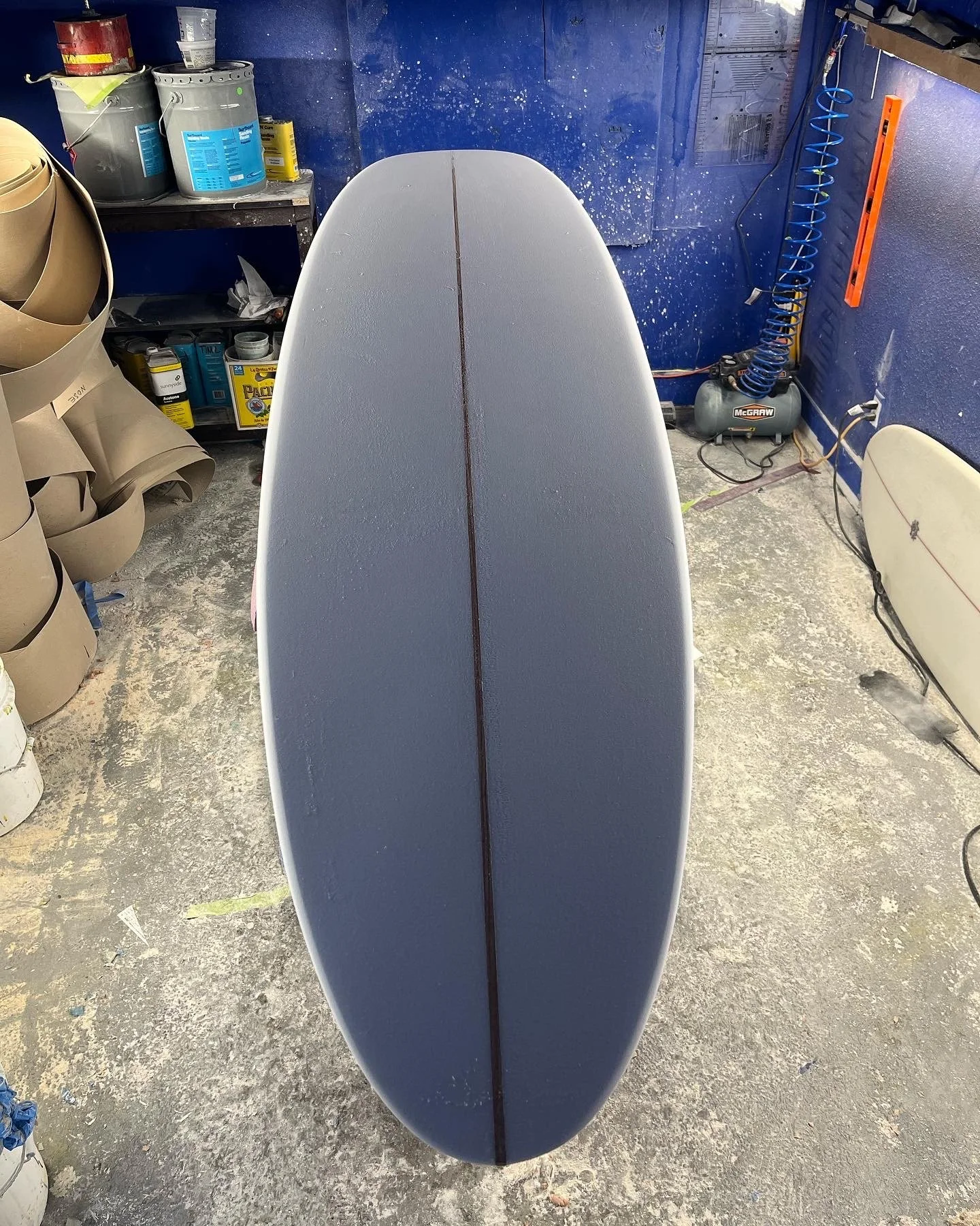 The image shows the nose of a surfboard with a visible stringer, standing on a concrete floor in a workshop or garage area. Surrounding the surfboard are various tools, paint cans, and equipment including a portable air compressor, rolls of cardboard