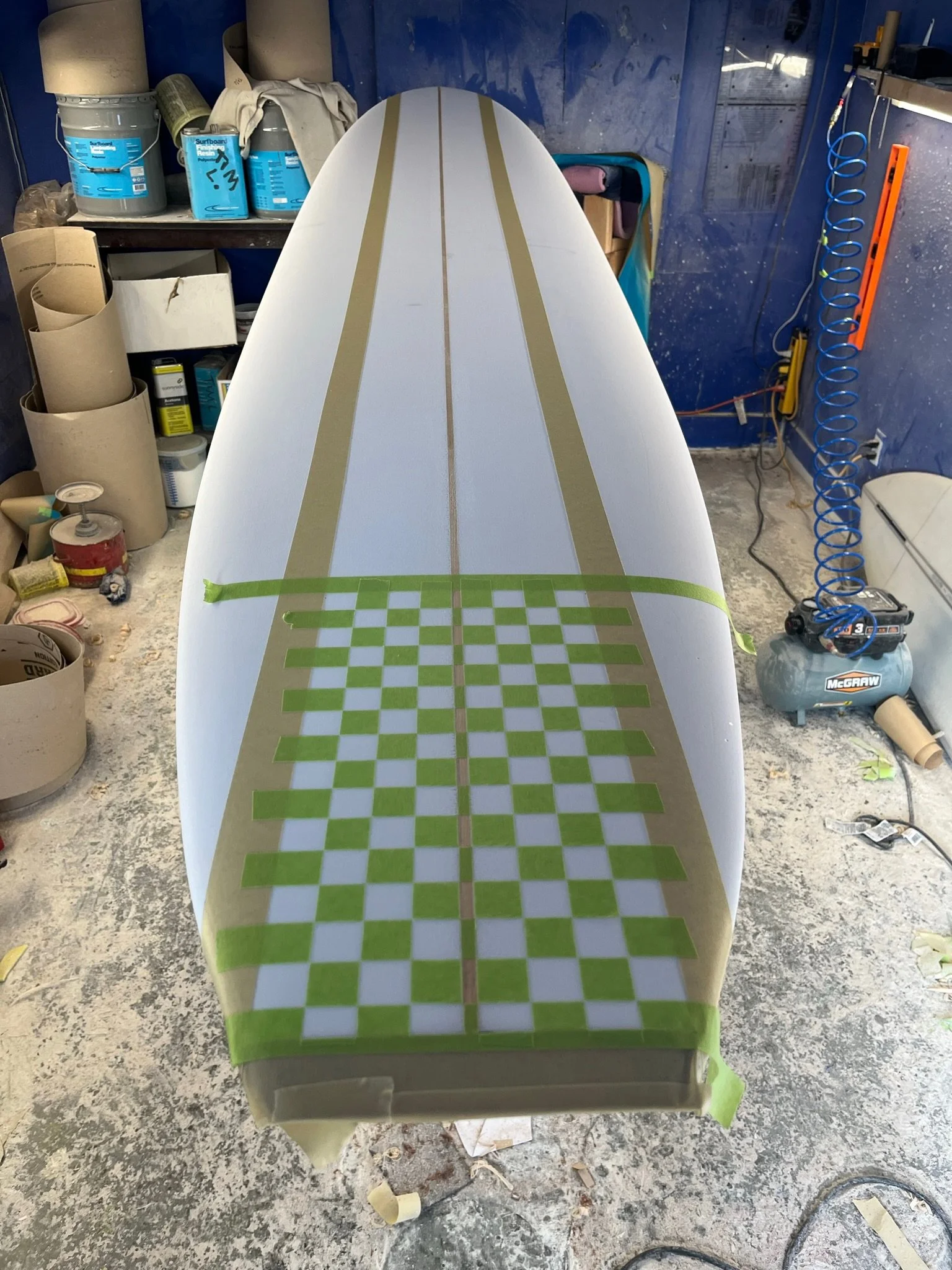 A boat hull in a workshop with masking tape and checkered pattern, surrounded by various tools and supplies, with blue walls and a cluttered workspace.